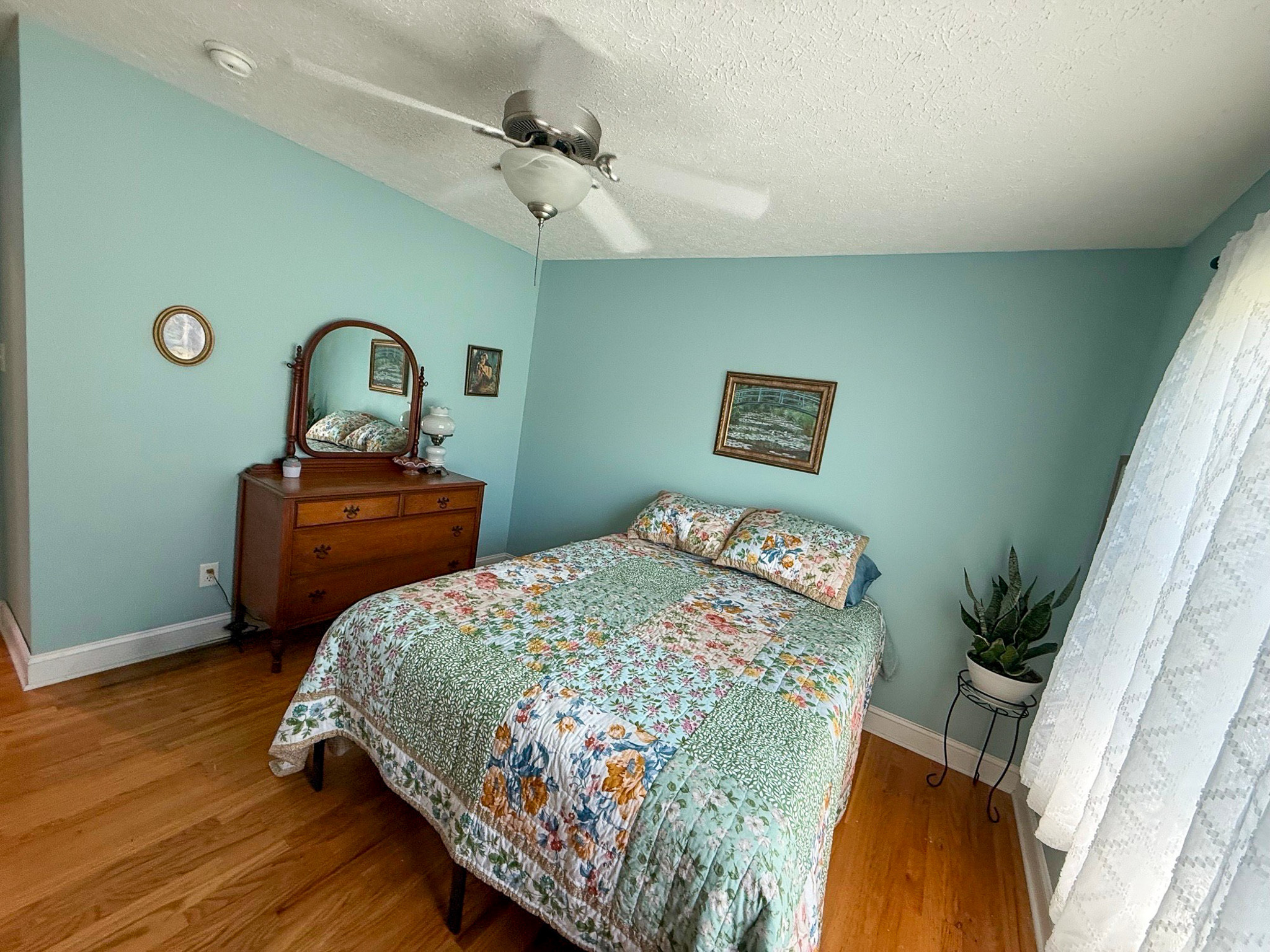 A bedroom with light green walls, a patterned quilt, a wooden dresser with a mirror, and a ceiling fan in the home of Group Home Providers Kim and Rick Duncan in Patrick Springs, Virginia.