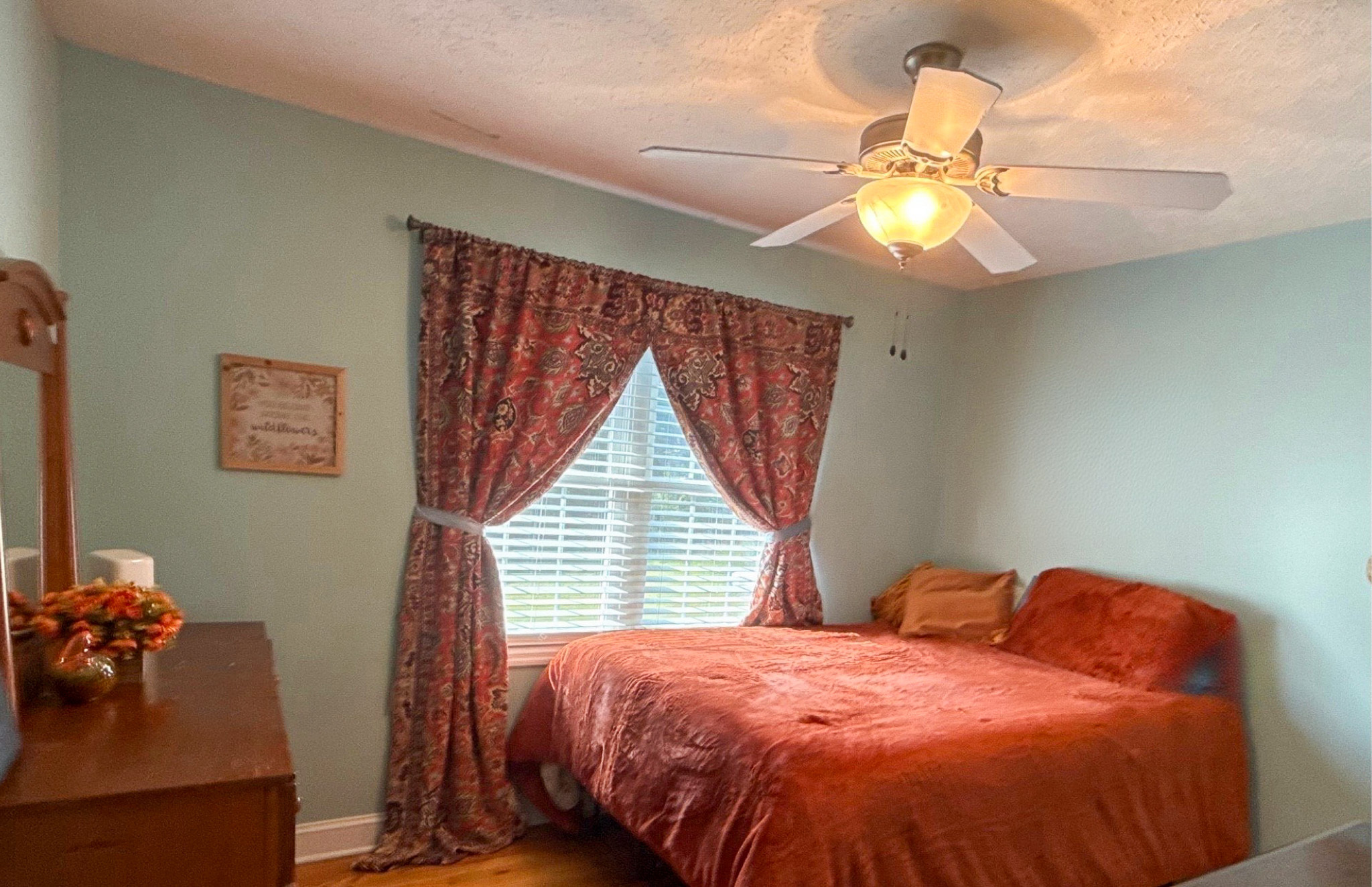 A bedroom with light green walls, a bed with rust-colored bedding, patterned curtains, a dresser, and a ceiling fan with a light in the home of Group Home Providers Kim and Rick Duncan in Patrick Springs, Virginia.