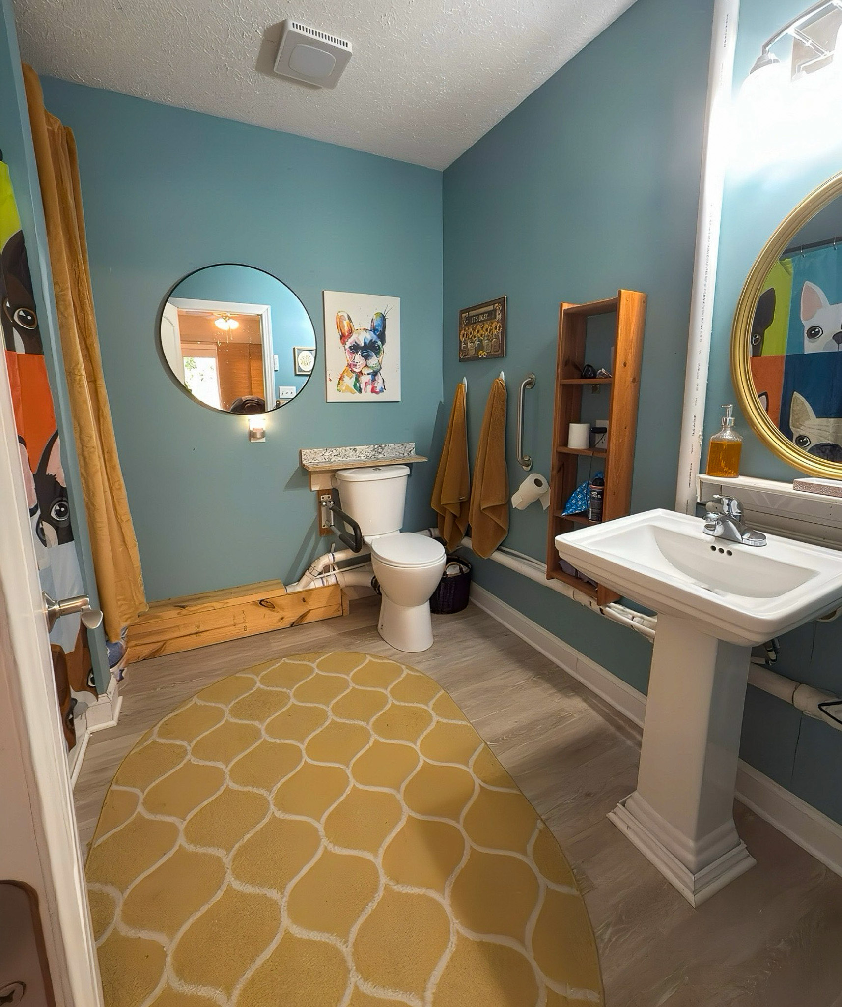 A bathroom with blue walls, a pedestal sink, a toilet, round mirrors, and yellow towels beside a patterned rug in the home of Group Home Providers Kim and Rick Duncan in Patrick Springs, Virginia.