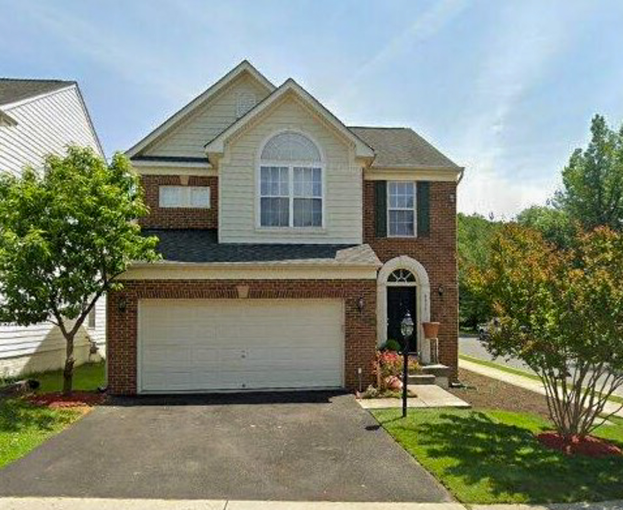 Two-story brick and siding suburban house with a double garage, front lawn, and trees on a sunny day belonging to Sponsored Residential provider Eglee Escorihuela.