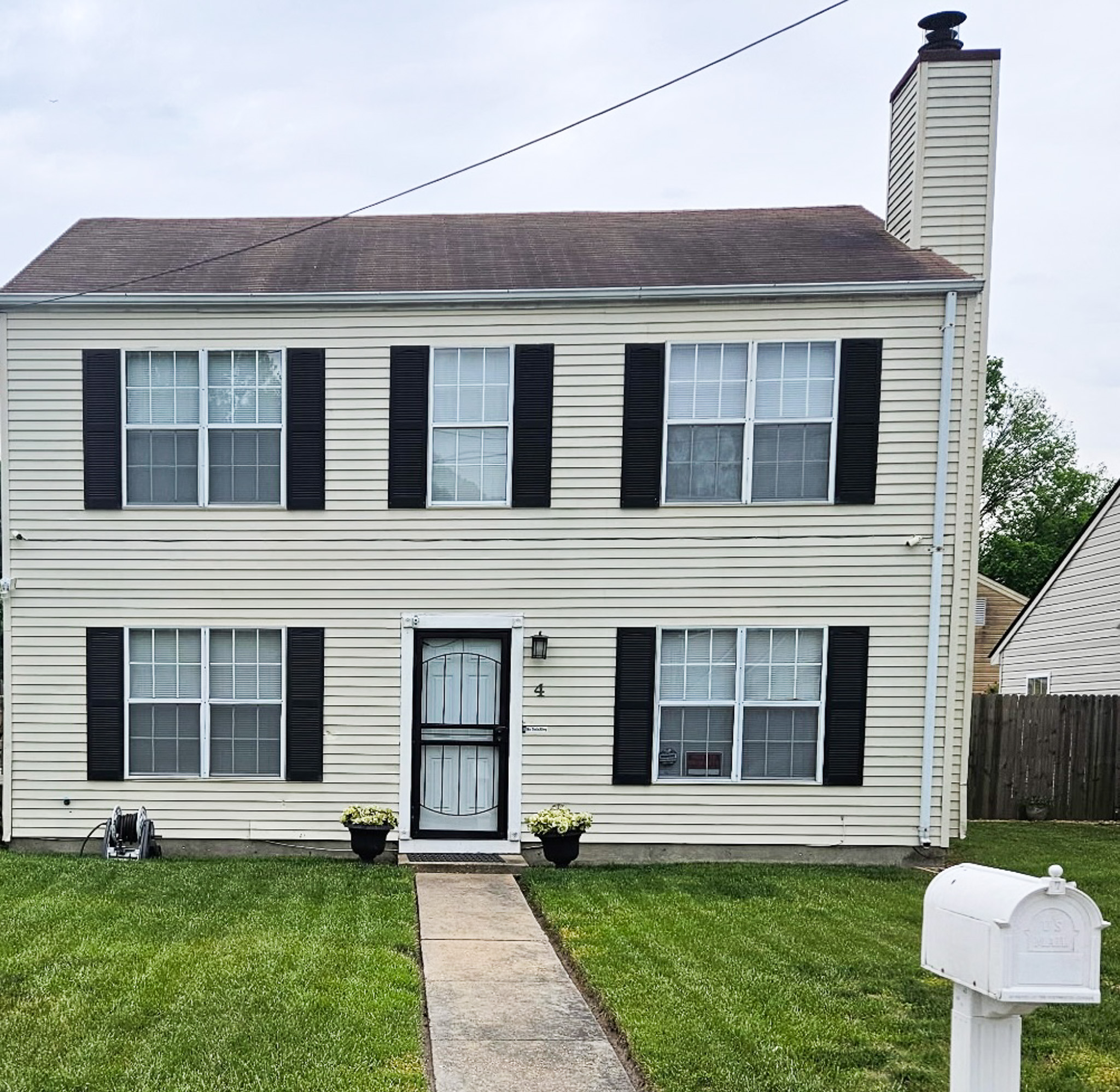 Two‑story beige house with black shutters, centered front door, small walkway across a green lawn, and a white mailbox in the foreground belonging to Sponsored Residential Provider Youlina Gamble in Hampton, Virginia.