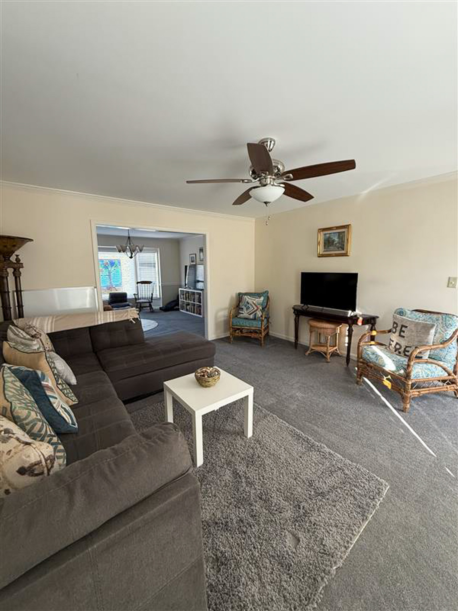 Living room with sectional sofa, ceiling fan, TV on a stand, wicker chairs, and neutral décor, opening into a dining area inside the home of Sponsored Residential Provider Gamu Gomo in Woodbridge, Virginia.
