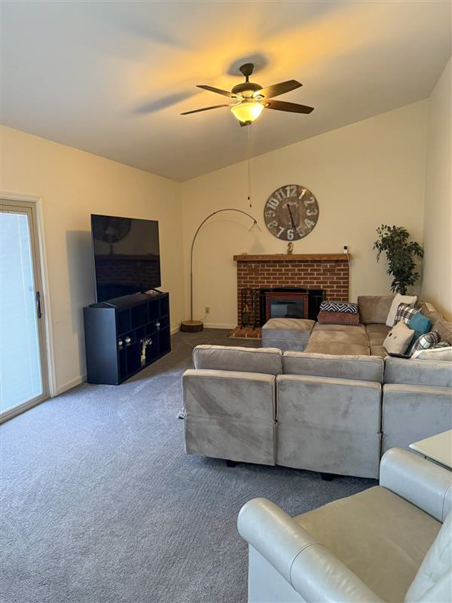 Living room with sectional sofa, TV stand, ceiling fan, brick fireplace, and sliding glass door leading outside inside the home of Sponsored Residential Provider Gamu Gomo in Woodbridge, Virginia.