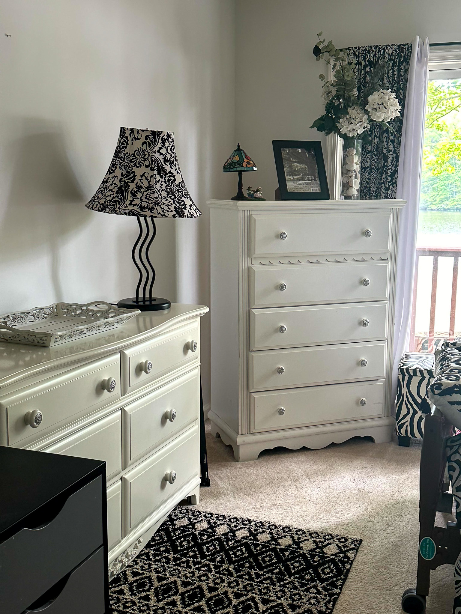 Bedroom corner with white dressers, a patterned lamp, floral decor, and a window with black‑and‑white curtains inside the home of Group Home Provider Ana Garcia in Spotsylvania, Virginia.