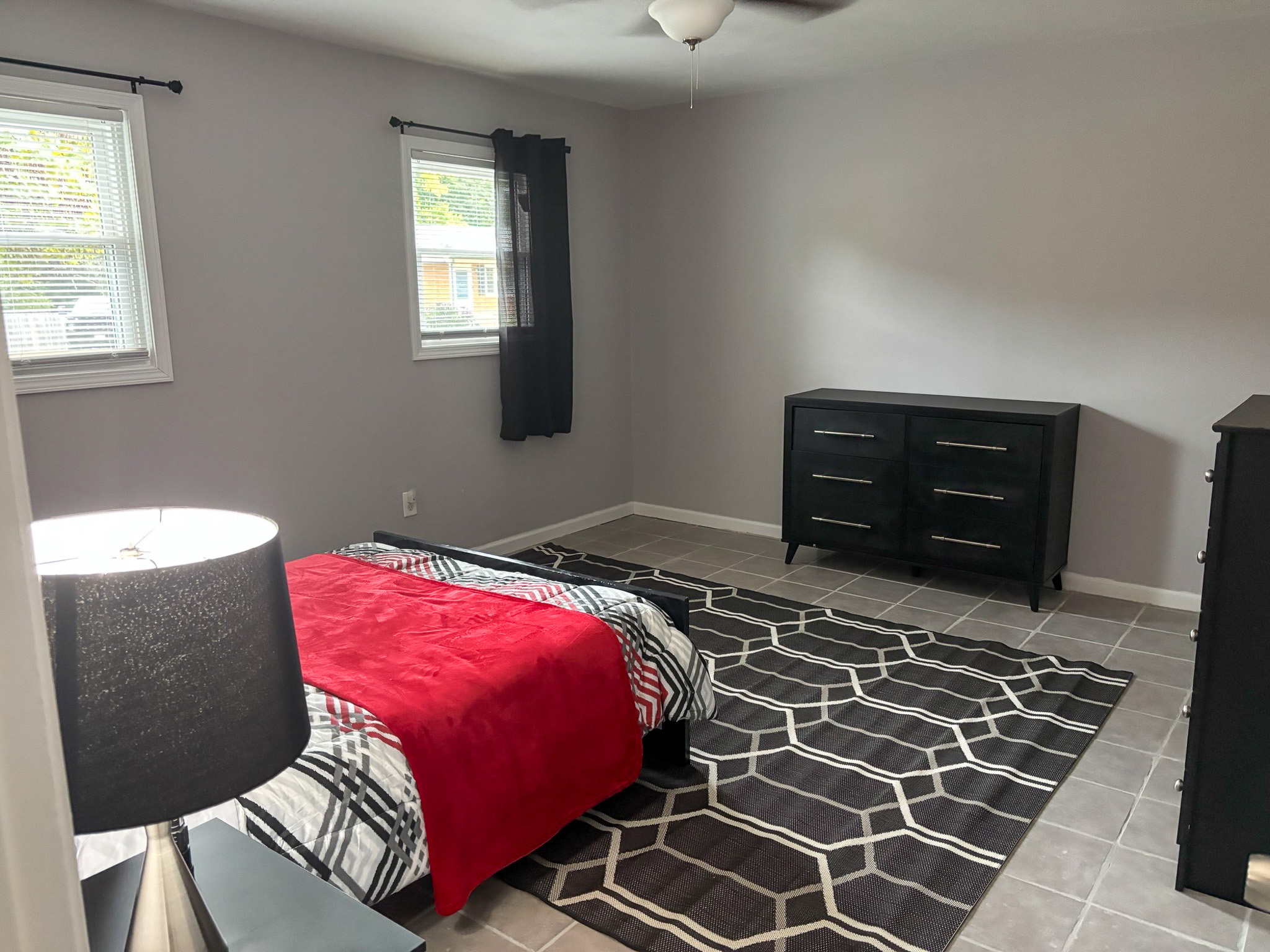 A spacious bedroom with a black bed frame, geometric rug, and matching black dresser under two windows with dark curtains inside the home of Sponsored Residential Providers Hadassah Hawks and Antonio Hernandez in Salem, Virginia.