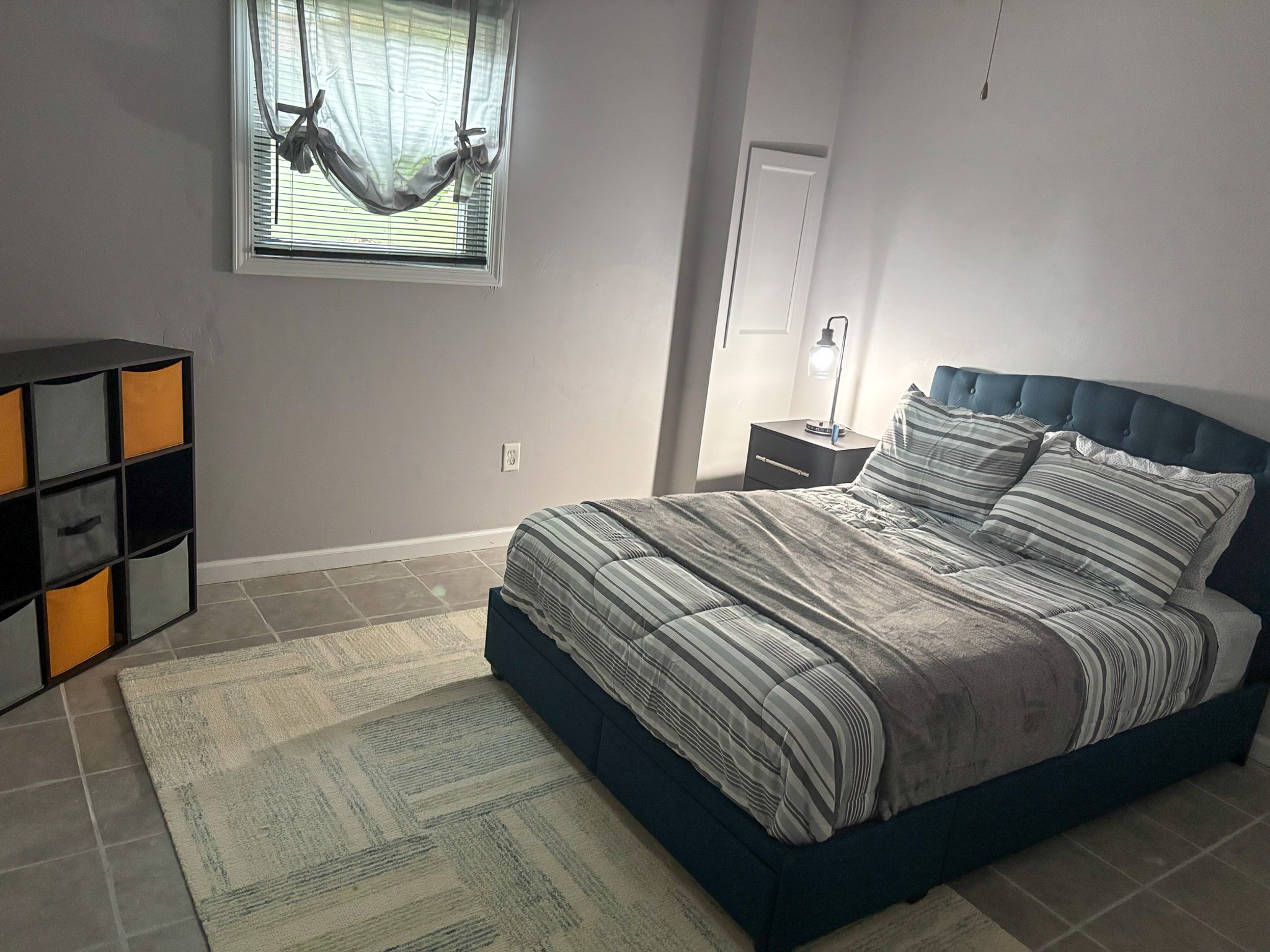 A bedroom with a neatly made striped bed, a small bedside lamp, a cube storage shelf, and a window with tied curtains inside the home of Sponsored Residential Providers Hadassah Hawks and Antonio Hernandez in Salem, Virginia.