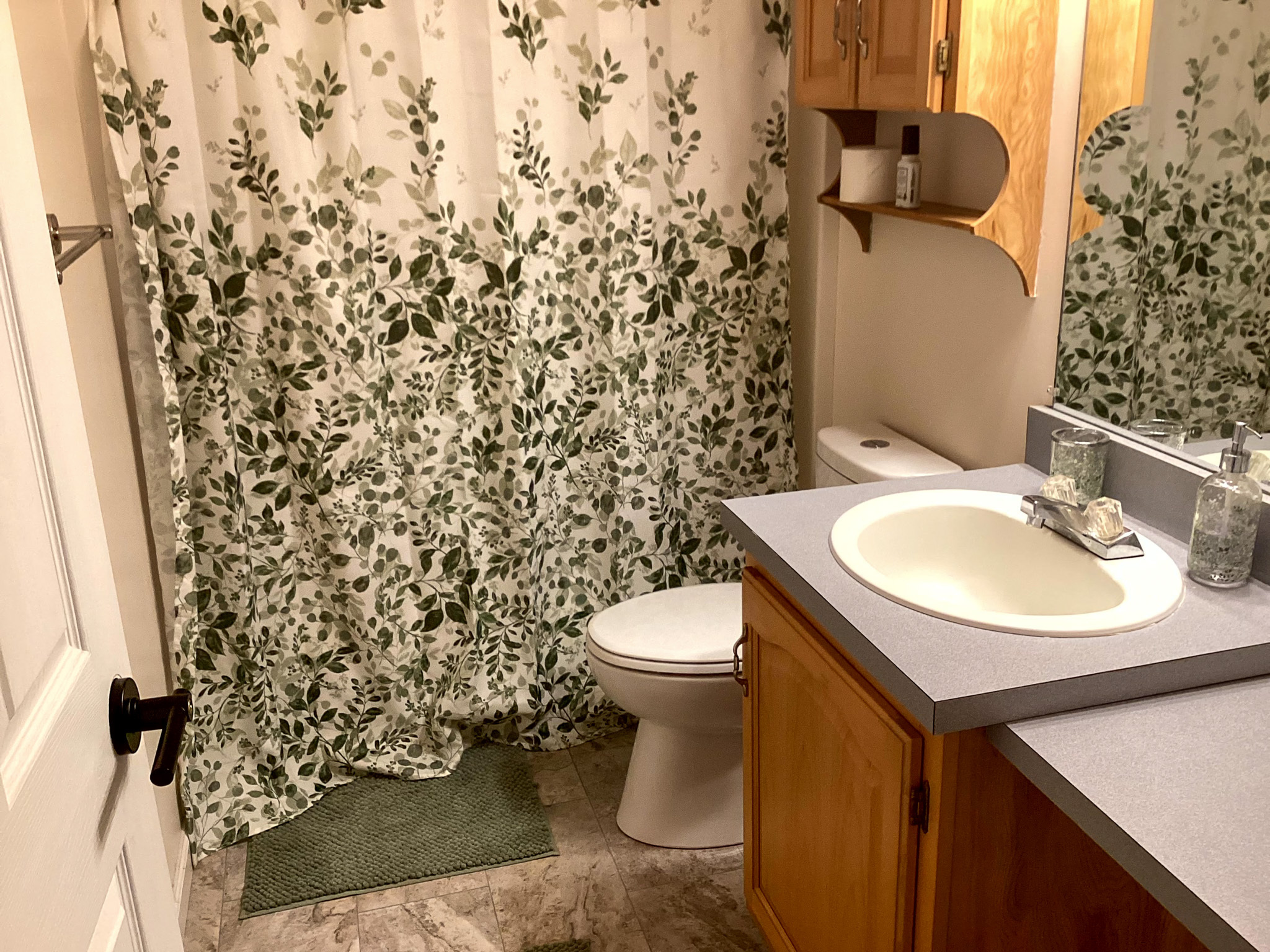 Bathroom with a leafy-pattern shower curtain, wood vanity, white sink, and toilet with a small green rug inside the home of Sponsored Residential Provider Kristen Hensley in Madison County, Virginia.