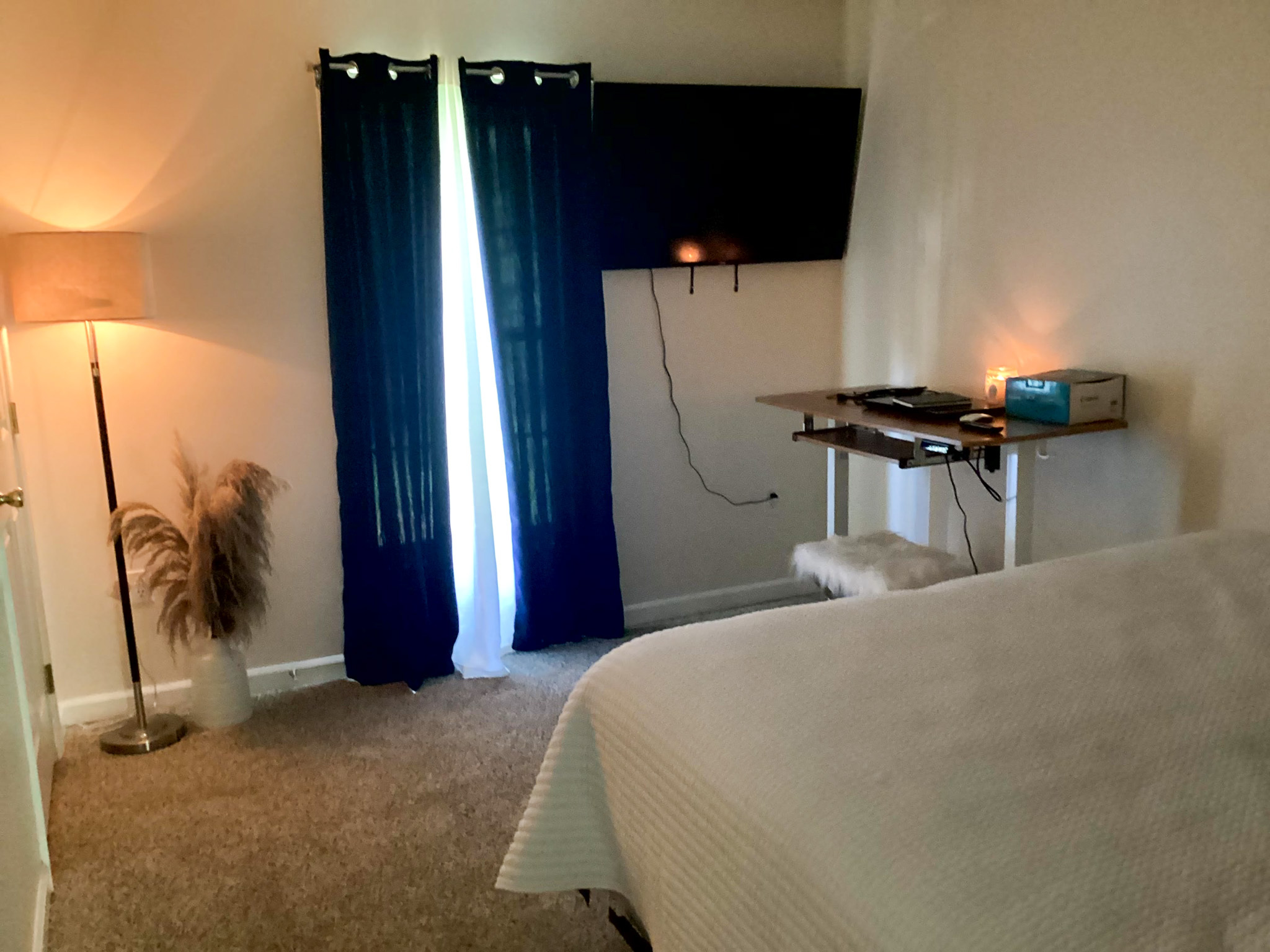 Bedroom with a bed in the foreground, dark curtains over a window, wall-mounted TV, standing lamp, and small desk with a candle inside the home of Sponsored Residential Provider Kristen Hensley in Madison County, Virginia.
