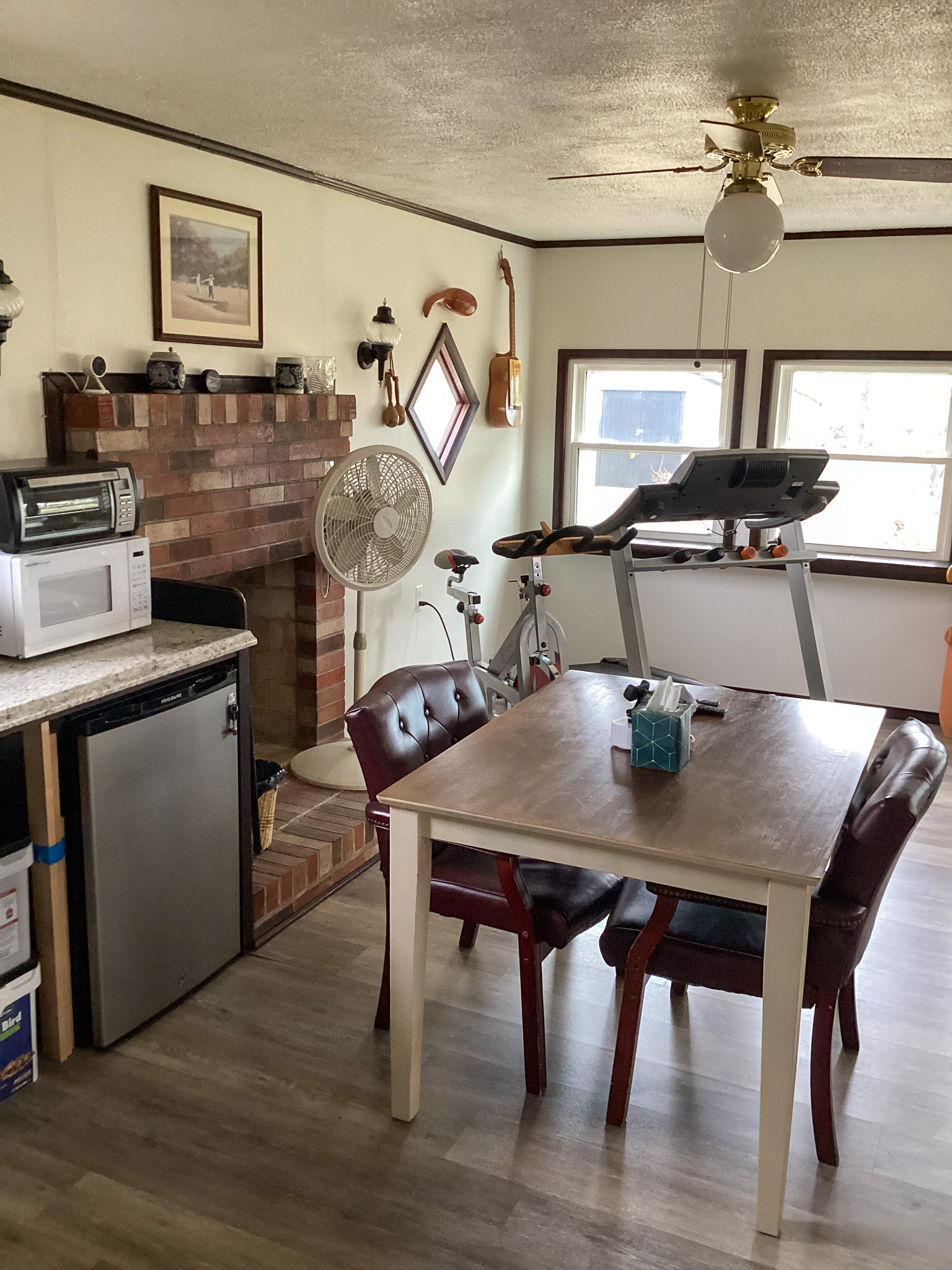 A small dining area with a wooden table and four chairs sits next to a brick fireplace, with a treadmill by the windows and various appliances on shelves to the left inside the home of Sponsored Residential Providers Teresa and Billy Herran in Woodbridge, Virginia.