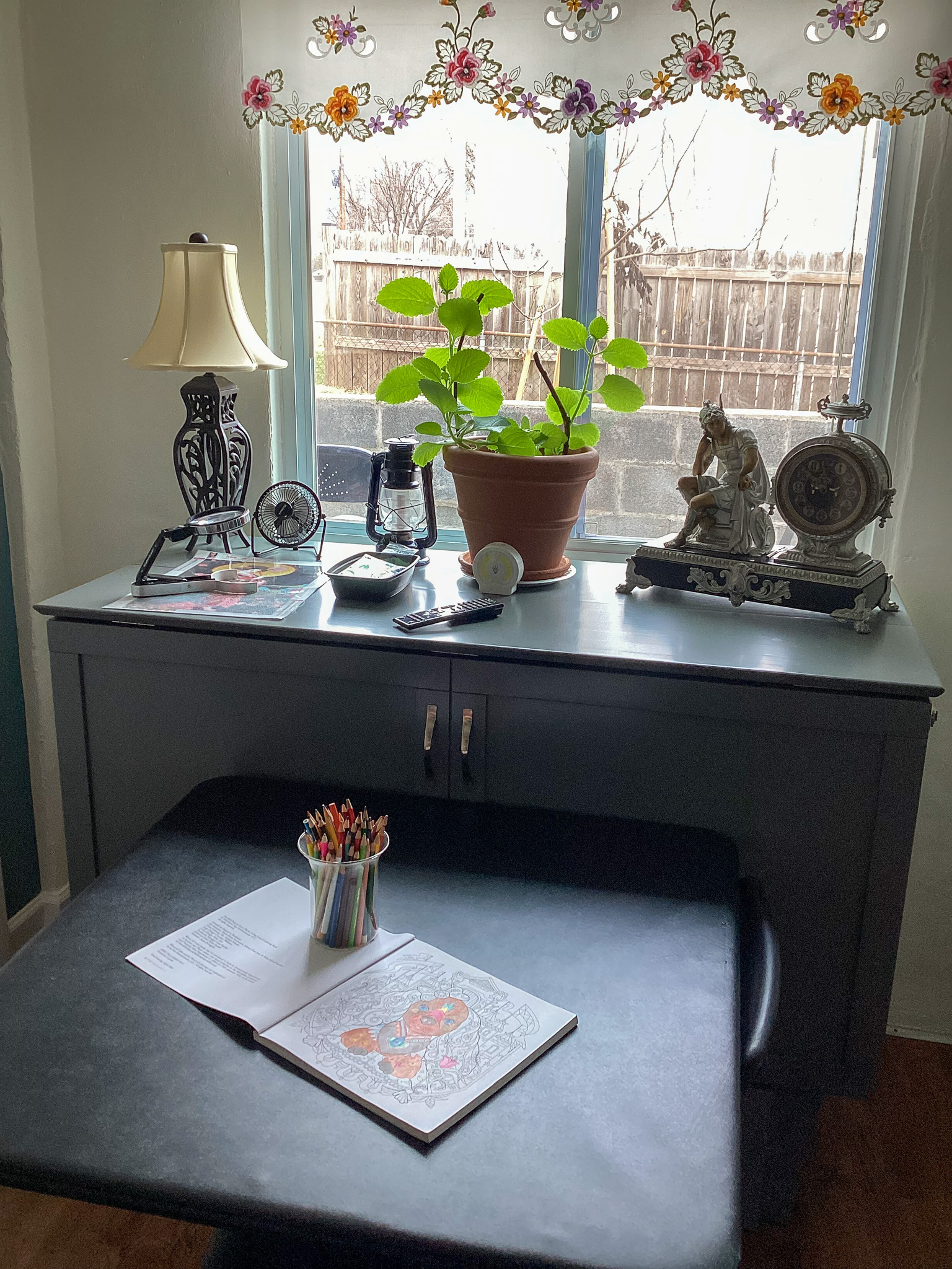 A small desk sits beneath a window with floral curtains, holding a potted plant, a lamp, decorative figurines, and various small items, with an open coloring book and a cup of crayons on a chair in front, inside the home of Sponsored Residential Providers Teresa and Billy Herran in Woodbridge, Virginia.