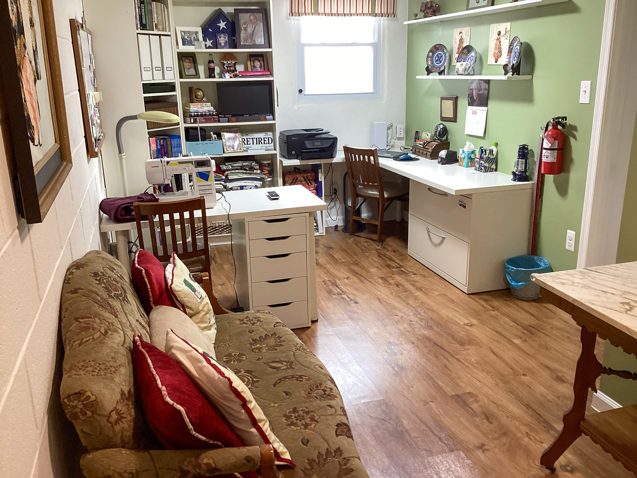 A cozy craft and workspace with a small sofa and pillows on the left, a central desk with drawers, shelves filled with supplies, and a second desk along a green accent wall, inside the home of Sponsored Residential Providers Teresa and Billy Herran in Woodbridge, Virginia.