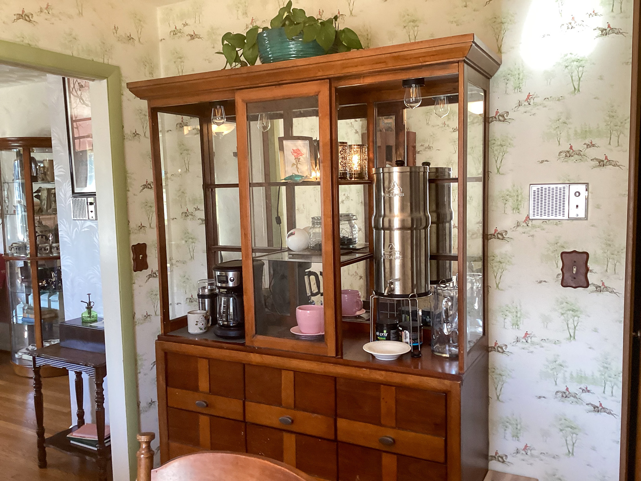 Wood china cabinet with glass doors displaying dishes and glassware, set against patterned wallpaper, with a glimpse into an adjoining room inside the home of Sponsored Residential Providers Bill and Heather Terry in Staunton, Virginia.