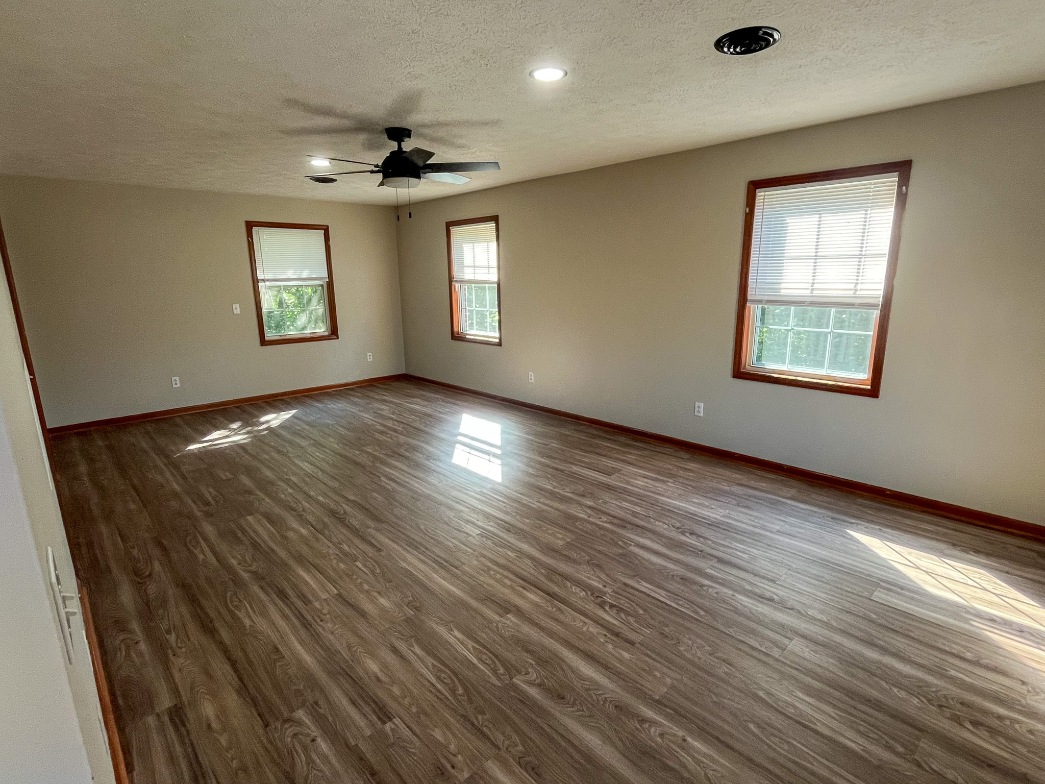 Large empty room with wood-look flooring, three windows, neutral walls, recessed lighting, and a ceiling fan  inside the home of Sponsored Residential Providers Penny Day and Steven Coleman in Hillsville, Virginia.
