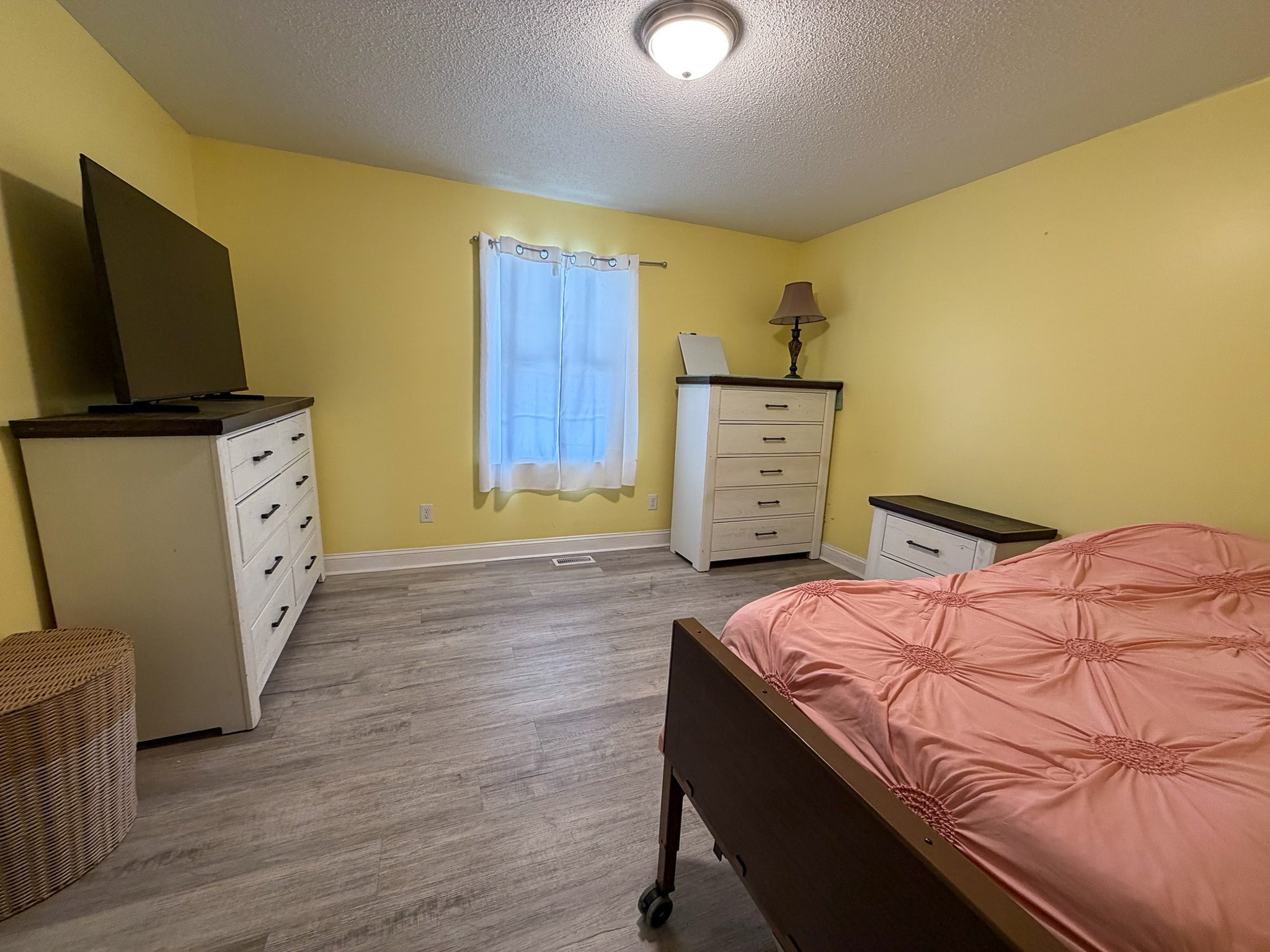 Bedroom with yellow walls, a bed with a pink comforter, light wood flooring, white dressers, a TV on a dresser, and a window with sheer curtains inside a Wall Residences Agency-Run Group Home.