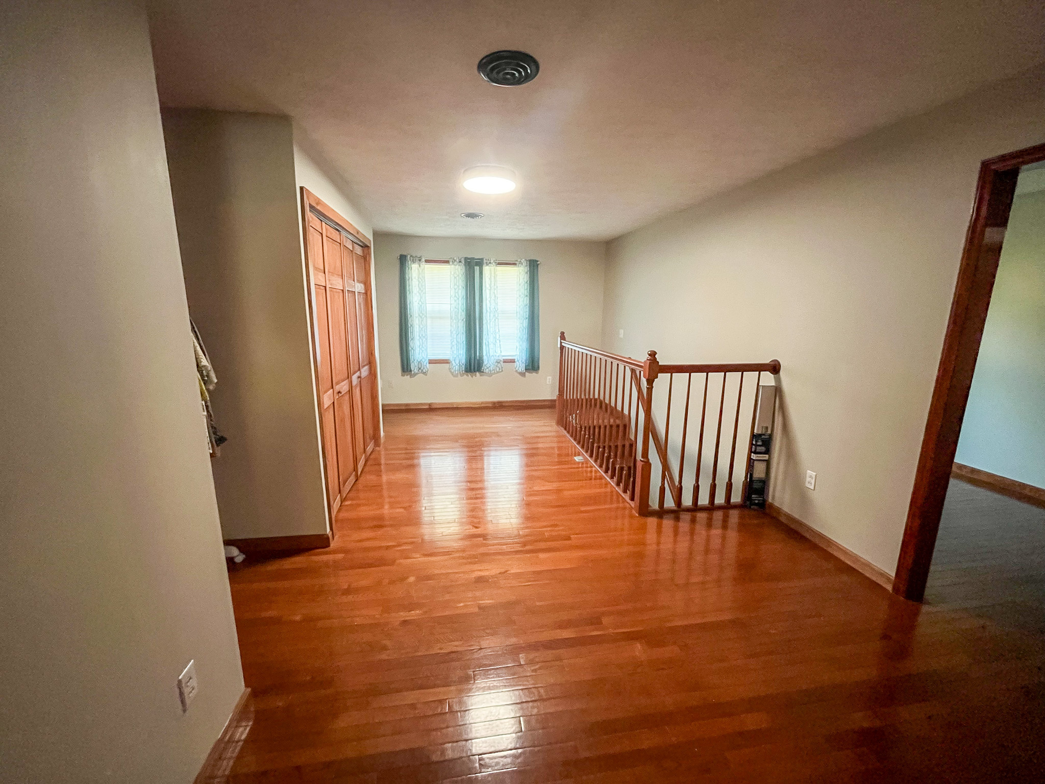 Hallway-style landing with hardwood floors, a window with blue curtains, wooden railing along a staircase, and double-door closets on one wall inside the home of Sponsored Residential Providers Penny Day and Steven Coleman in Hillsville, Virginia.