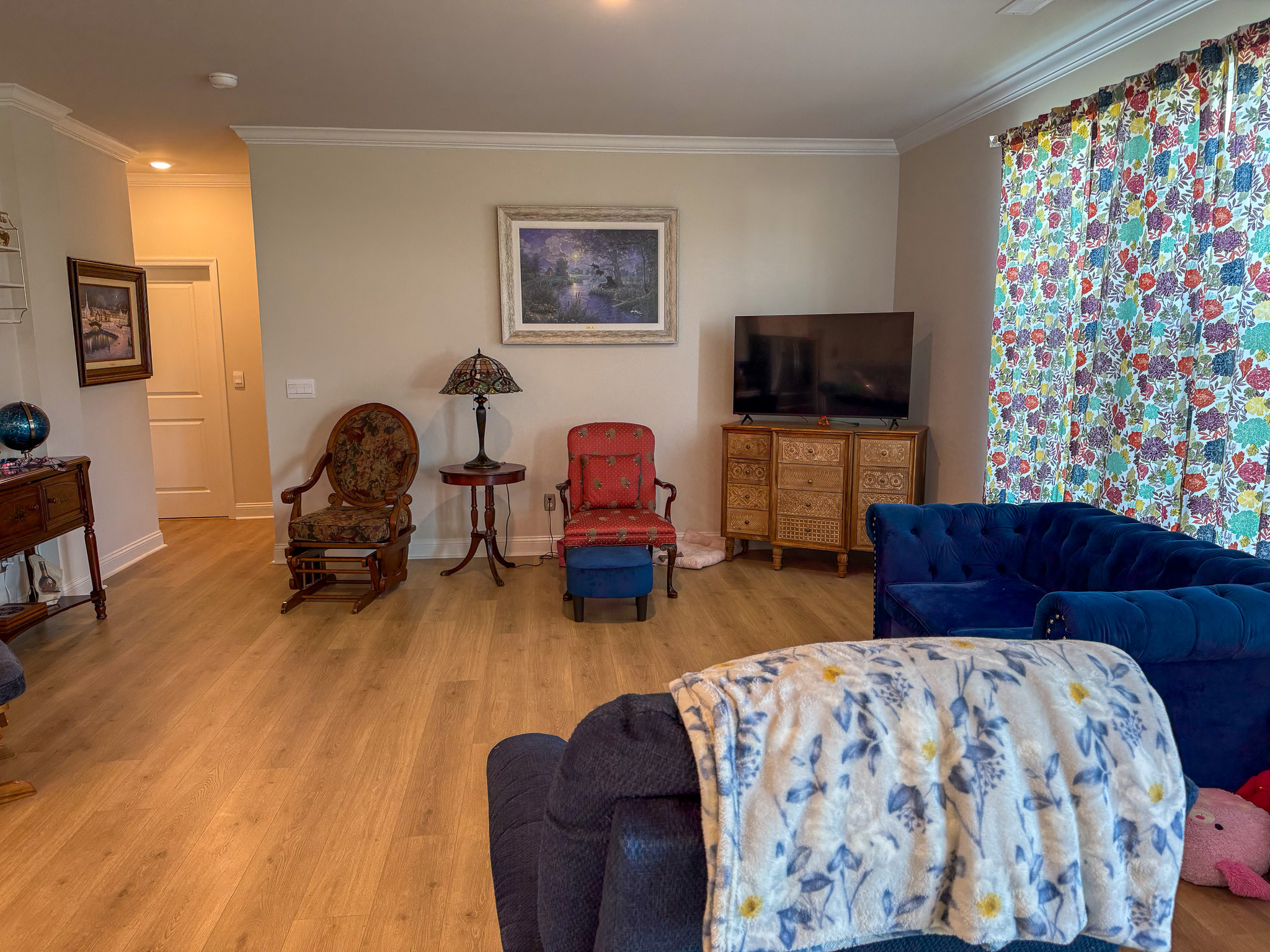 Living room with seating chairs, TV on a cabinet, wood flooring, and colorful curtains inside the home of Group Home provider Traci Burch in Staurts Draft, VA.