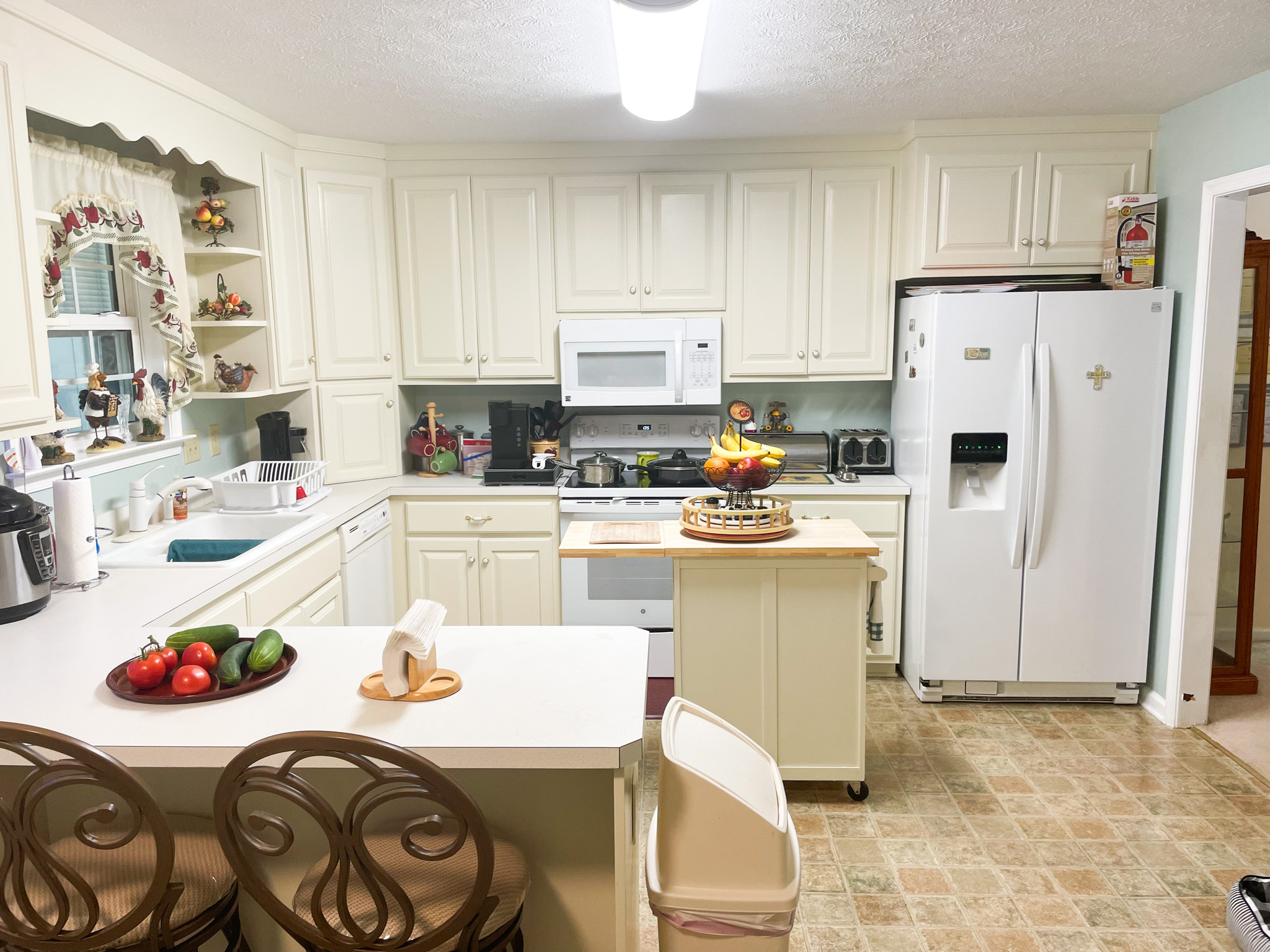 Bright kitchen with cream cabinets, white appliances, a central island with a fruit stand, and a breakfast bar with two metal stools  inside the home of Sponsored Residential Provider Kellie Lynch in Gloucester, Virginia.