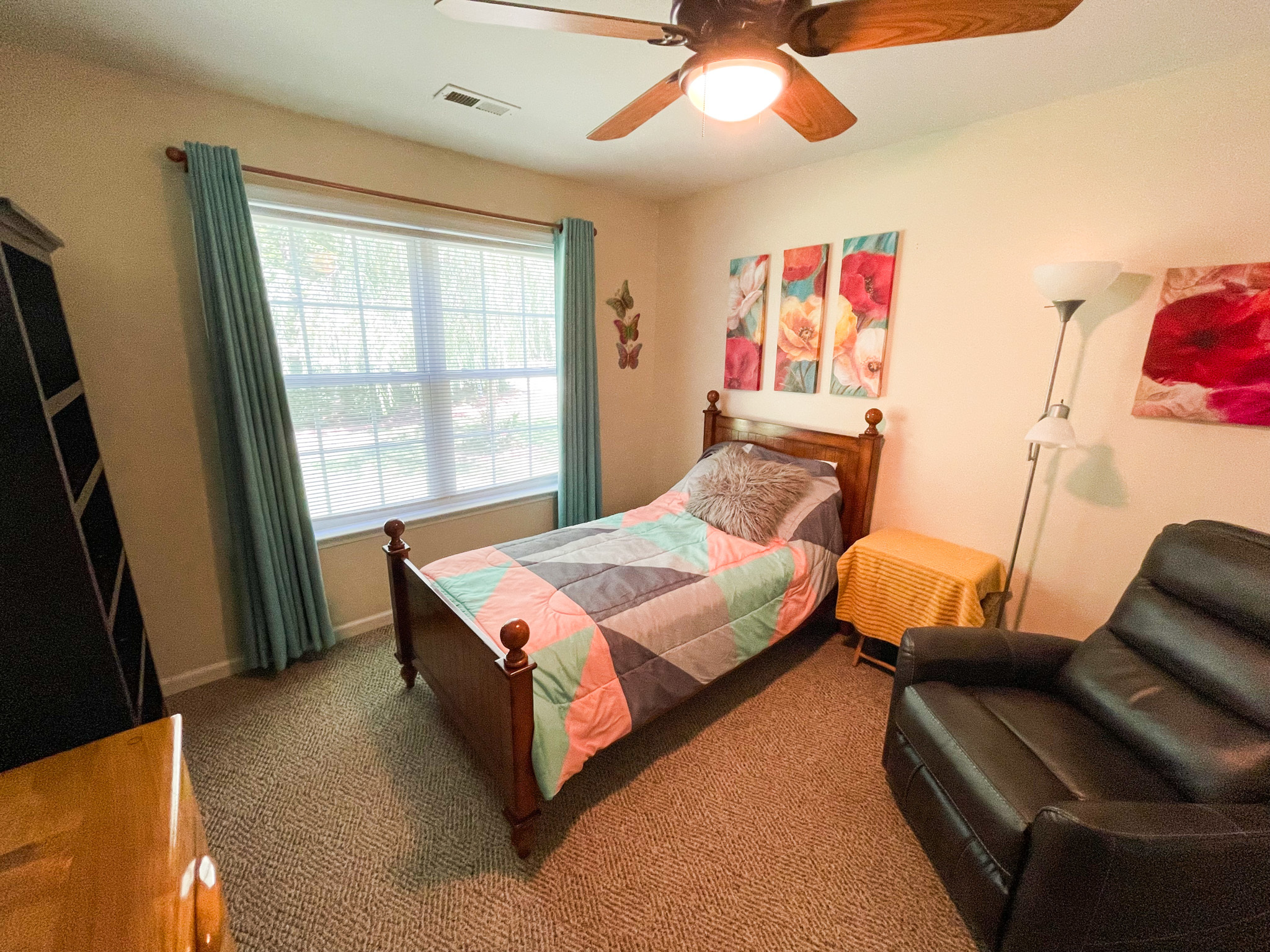Bedroom with a wooden bed frame, colorful patchwork quilt, large window with teal curtains, a recliner, and floral artwork on the wall inside the home of Sponsored Residential Provider Kellie Lynch in Gloucester, Virginia.