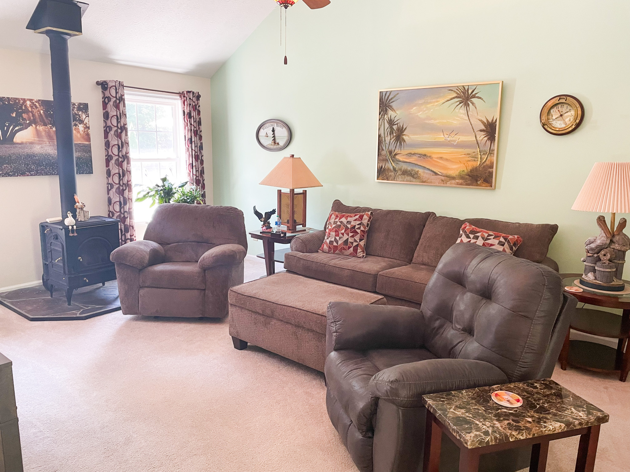 Living room with a brown sofa, matching recliners, a corner wood stove, and beach‑themed wall art on light green walls inside the home of Sponsored Residential Provider Kellie Lynch in Gloucester, Virginia.