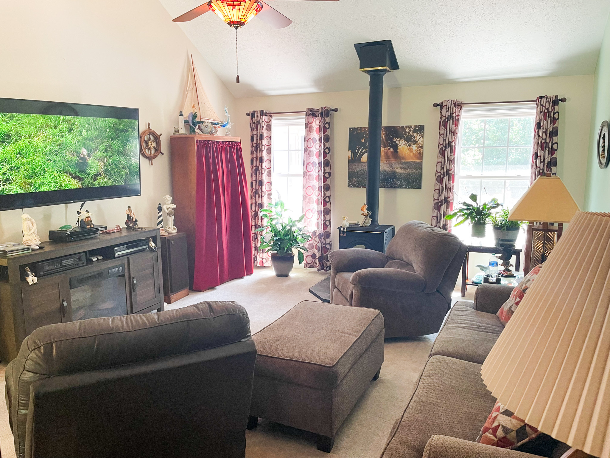 Living room with brown upholstered seating, a wood stove between two windows, and a large TV above a console on a light-colored wall  inside the home of Sponsored Residential Provider Kellie Lynch in Gloucester, Virginia.