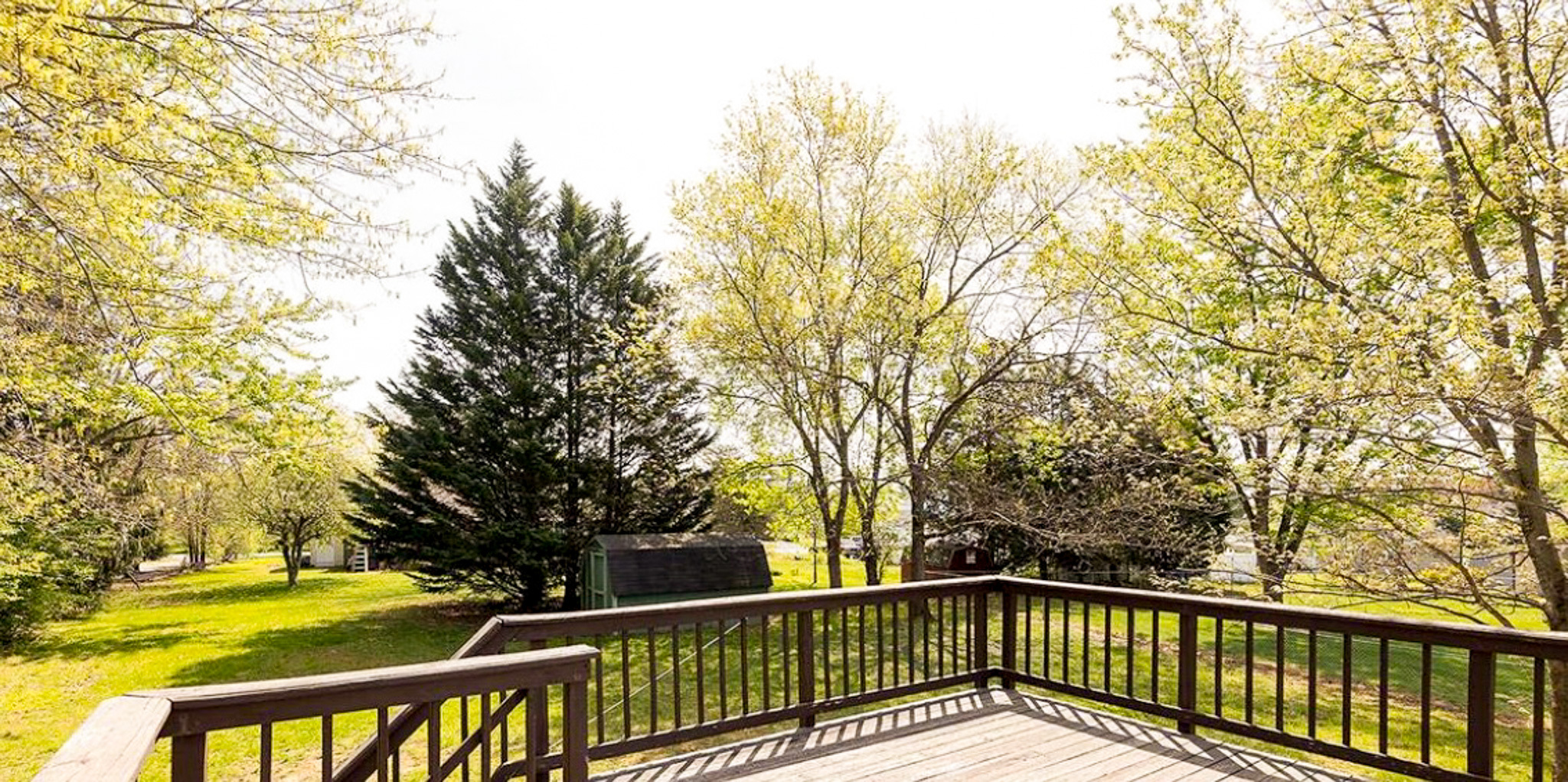 Wooden deck with railing overlooking a sunny backyard with mature trees and a small shed outside the home of Sponsored Residential Providers Daynesha and Karen Wilson in Stephens City, Virginia.