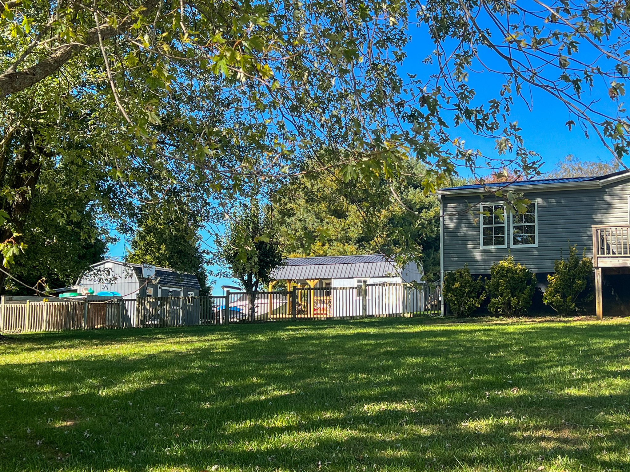 A large grassy yard shaded by a tree, with a gray house on the right and several outbuildings and fenced areas in the background at the home of Sponsored Residential Provider Christina Bedsaul in Hillsville, Virginia.