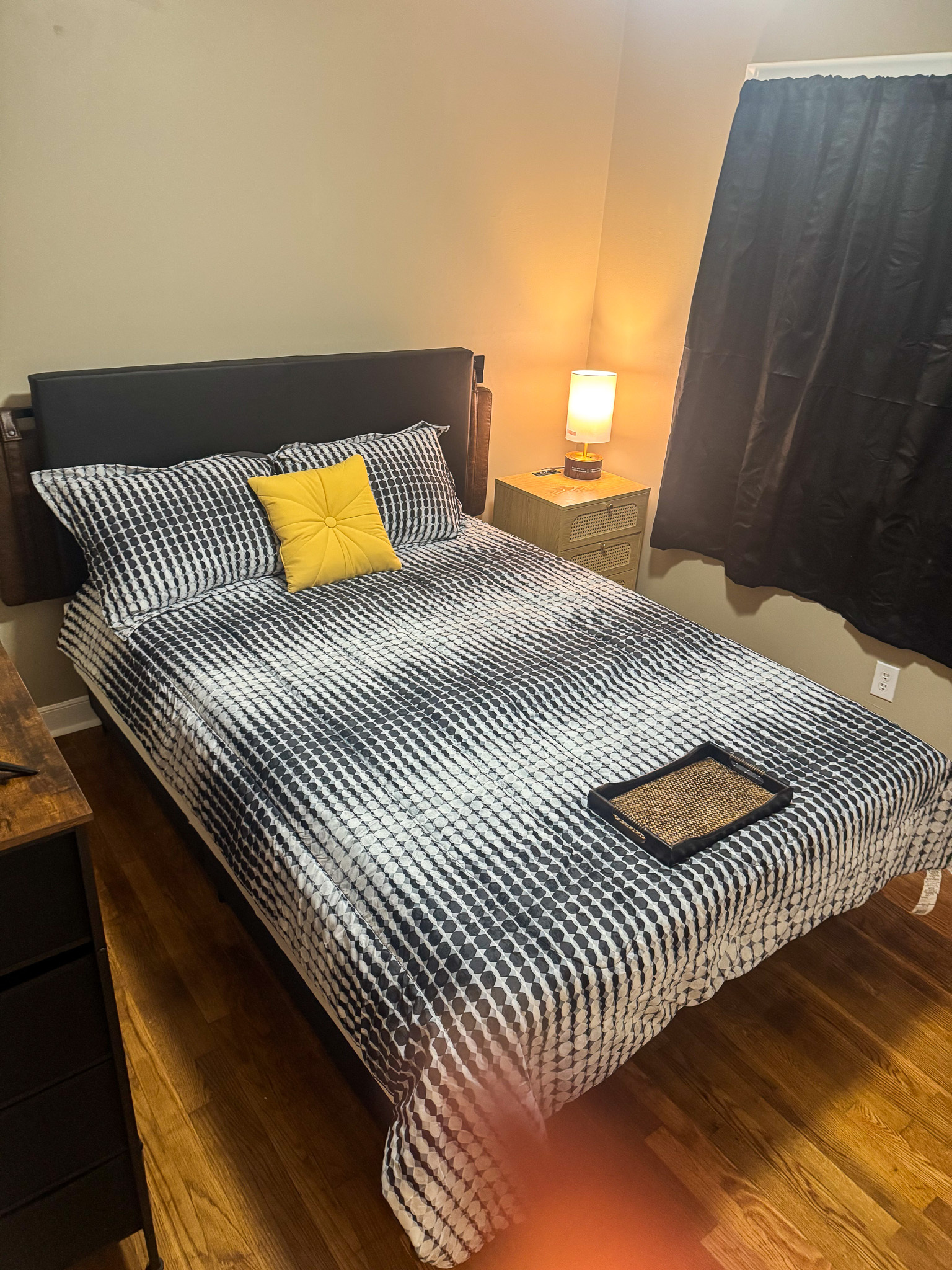 A bedroom with a bed dressed in a black‑and‑white patterned comforter, a yellow accent pillow, a bedside table with a lit lamp, dark curtains, and hardwood floors inside the home of Sponsored Residential Provider Selena Banks in Chesterfield, Virginia.