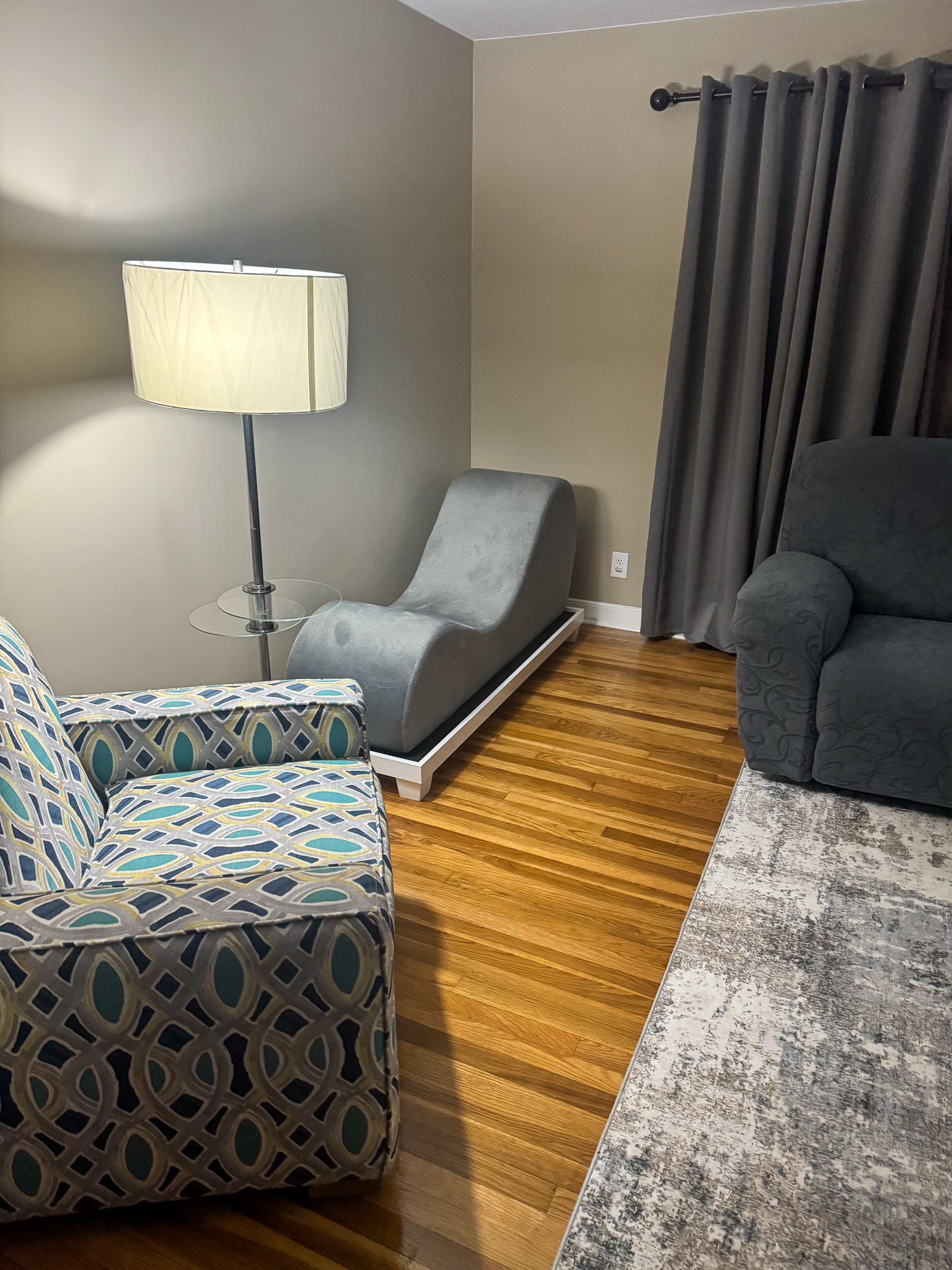 A living room corner with a patterned armchair, a floor lamp, a gray lounge chair, thick gray curtains, a dark armchair, and a large rug on hardwood floors inside the home of Sponsored Residential Provider Selena Banks in Chesterfield, Virginia.