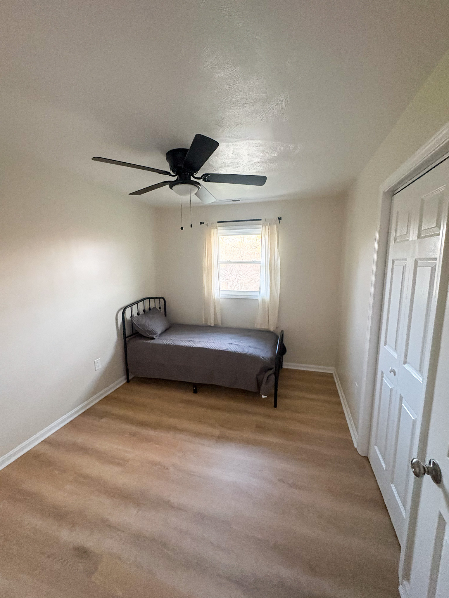 Simple bedroom with a single bed, light wood flooring, ceiling fan, small window with sheer curtains, and a closet door inside the home of sponsored Residential Provider Destiny Branscome in New River Valley, VA.