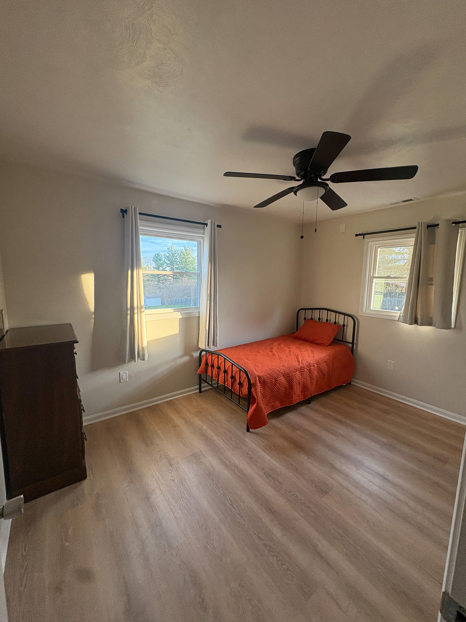Bedroom with a single bed featuring an orange blanket, light wood flooring, ceiling fan, two windows with curtains, and a small dresser inside the home of sponsored Residential Provider Destiny Branscome in New River Valley, VA.