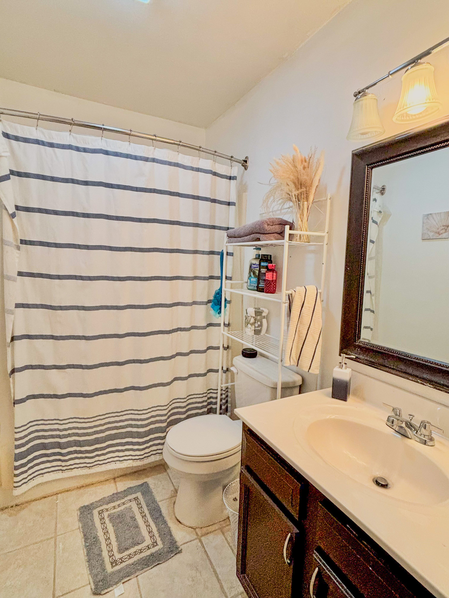 Bright bathroom with striped shower curtain, toilet, vanity sink, mirror, and storage shelf inside the home of Sponsored Residential Provider Lana Cyzicks in Staunton, Virginia.