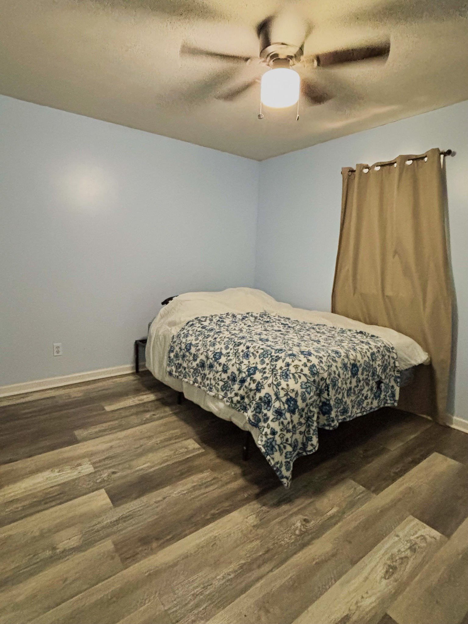 Light blue bedroom with a full‑size bed, patterned bedding, ceiling fan, and wood‑look flooring inside the home of Sponsored Residential Provider Lana Cyzicks in Staunton, Virginia.