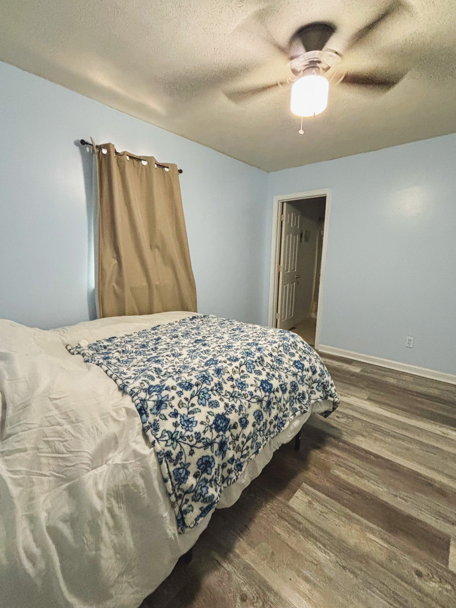 Bedroom with a full‑size bed, light blue walls, gray wood‑look flooring, ceiling fan, and window with curtain inside the home of Sponsored Residential Provider Lana Cyzicks in Staunton, Virginia.