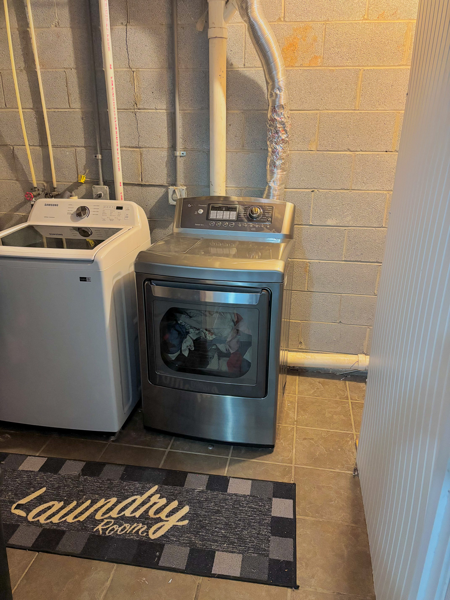Basement laundry room with washer, dryer, exposed pipes, and tiled floor inside the home of Sponsored Residential Provider Lana Cyzicks in Staunton, Virginia.