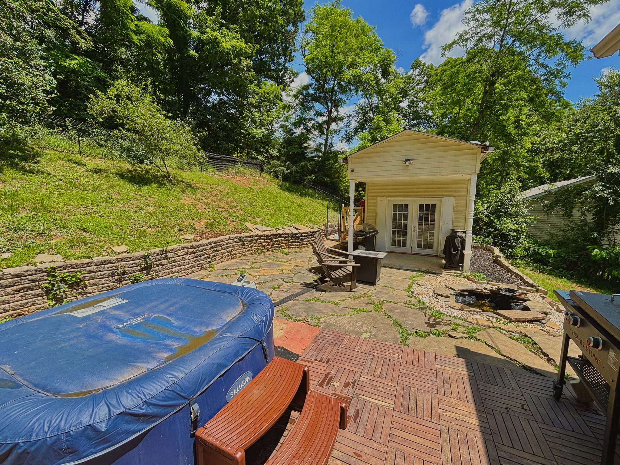 Backyard patio with hot tub, fire pit seating, stone pavers, grill, and small outbuilding surrounded by trees inside the home of Sponsored Residential Provider Lana Cyzicks in Staunton, Virginia.