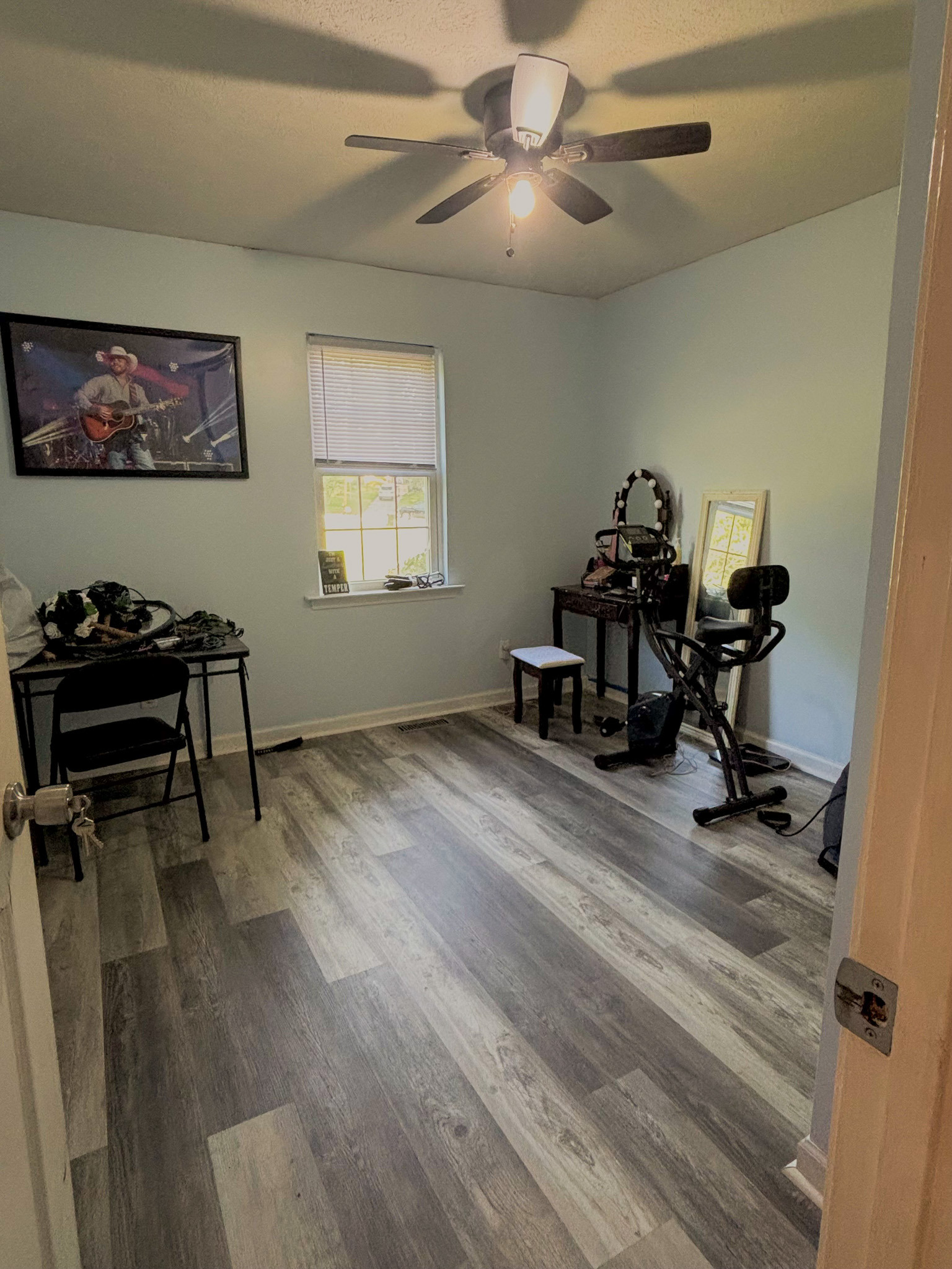 Empty bedroom with gray wood‑look flooring, ceiling fan, window, desk, and exercise bike inside the home of Sponsored Residential Provider Lana Cyzicks in Staunton, Virginia.