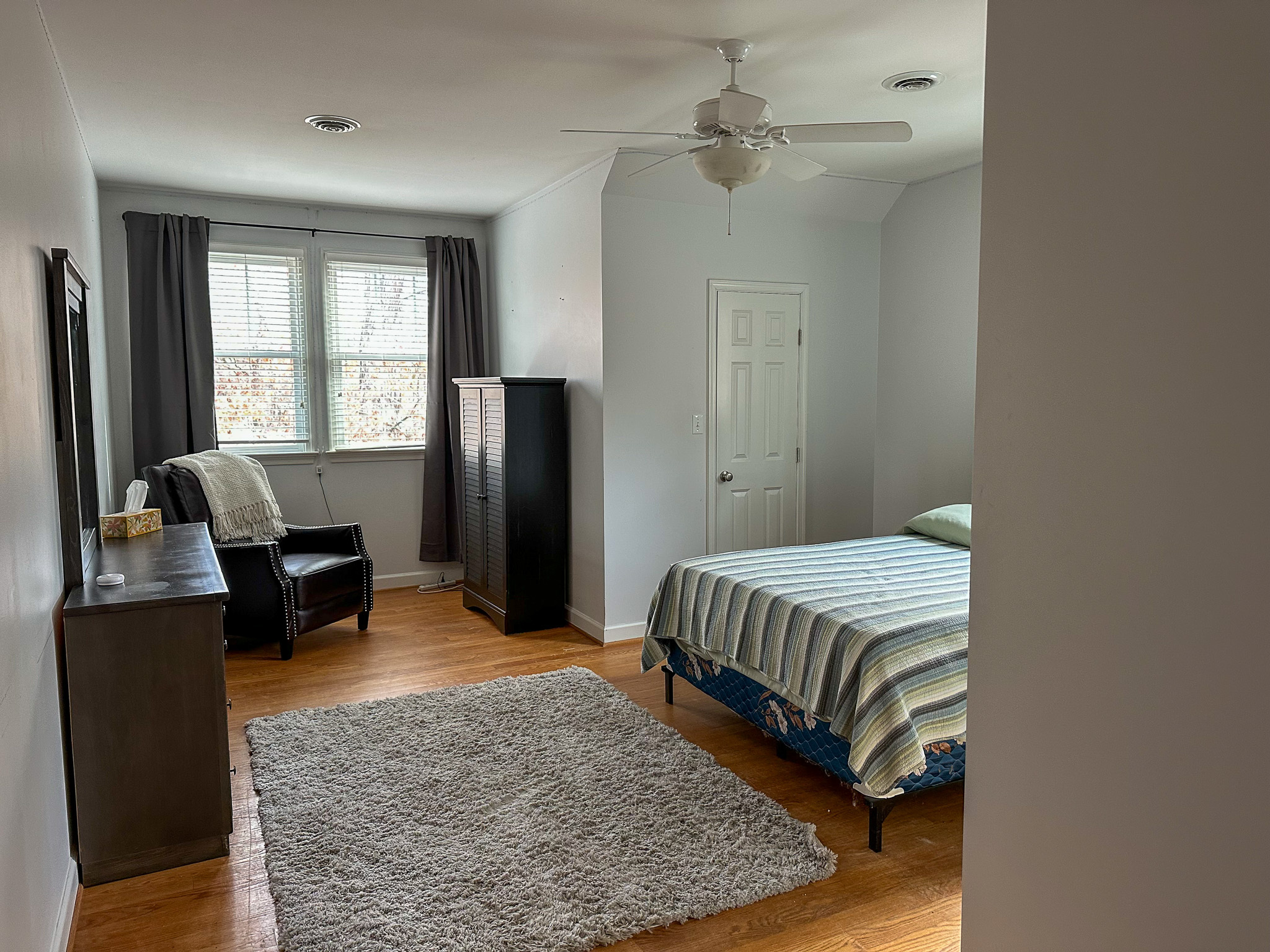 Bright bedroom with a neatly made bed, dresser, armchair by two large windows, and a light gray rug on a wood floor  inside the home of Group Home Providers Felicia and Jason Martin in Amherst, Virginia.