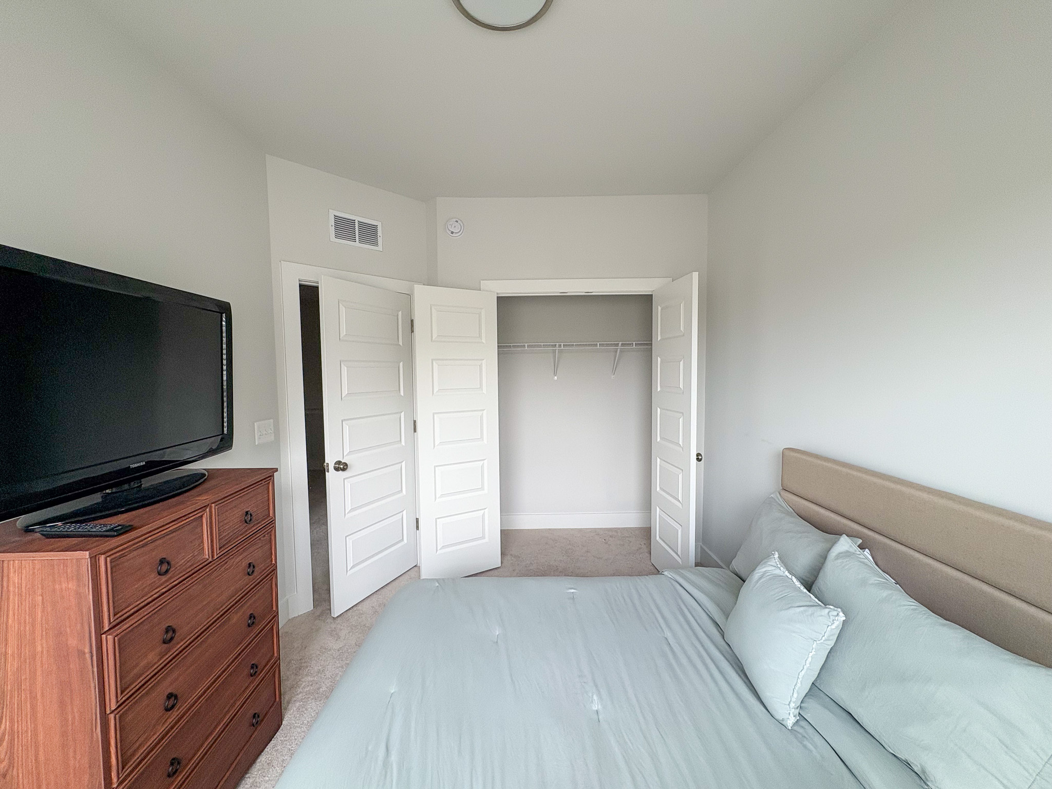 A bright bedroom with a neatly made bed, a wooden dresser with a large TV, white double doors leading to a closet, and soft natural light illuminating the space inside the home of Sponsored Residential Providers Tania and LeSean Matthews in Glen Allen, Virginia.