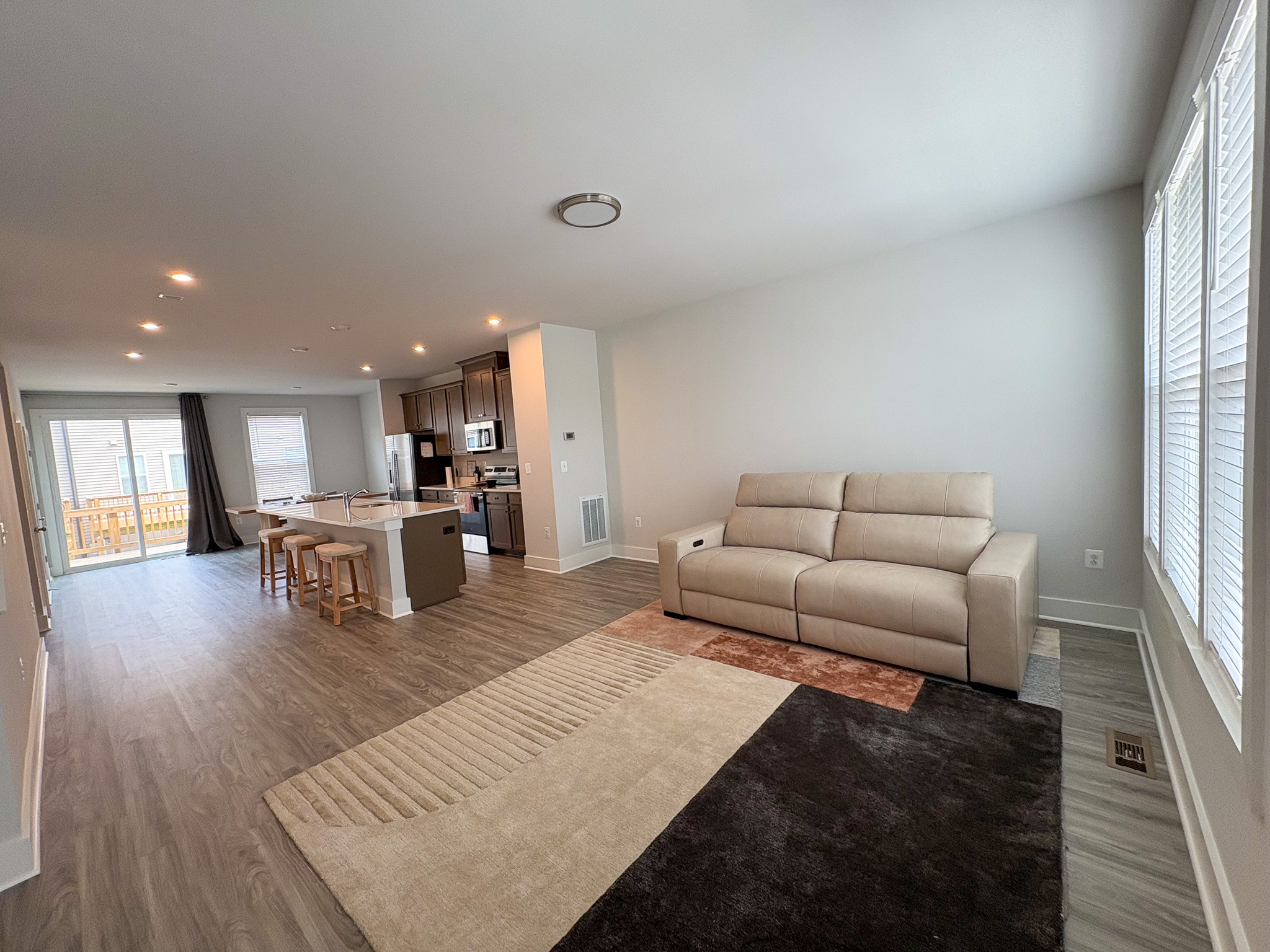 An open‑concept living space with a beige sofa on a two‑tone rug, wood‑look flooring, large windows, and a view into a kitchen and dining area with an island and sliding glass doors inside the home of Sponsored Residential Providers Tania and LeSean Matthews in Glen Allen, Virginia.