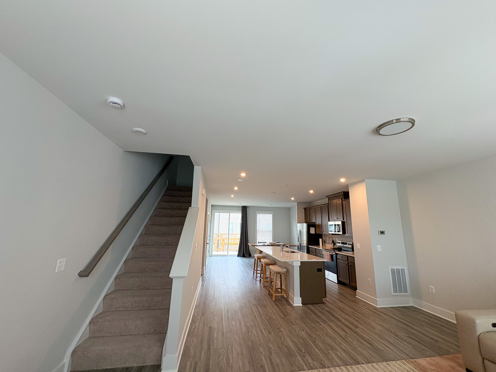 An open living area with a carpeted staircase on the left, wood‑look flooring, a large kitchen island with stools, and sliding doors leading to a bright outdoor space inside the home of Sponsored Residential Providers Tania and LeSean Matthews in Glen Allen, Virginia.