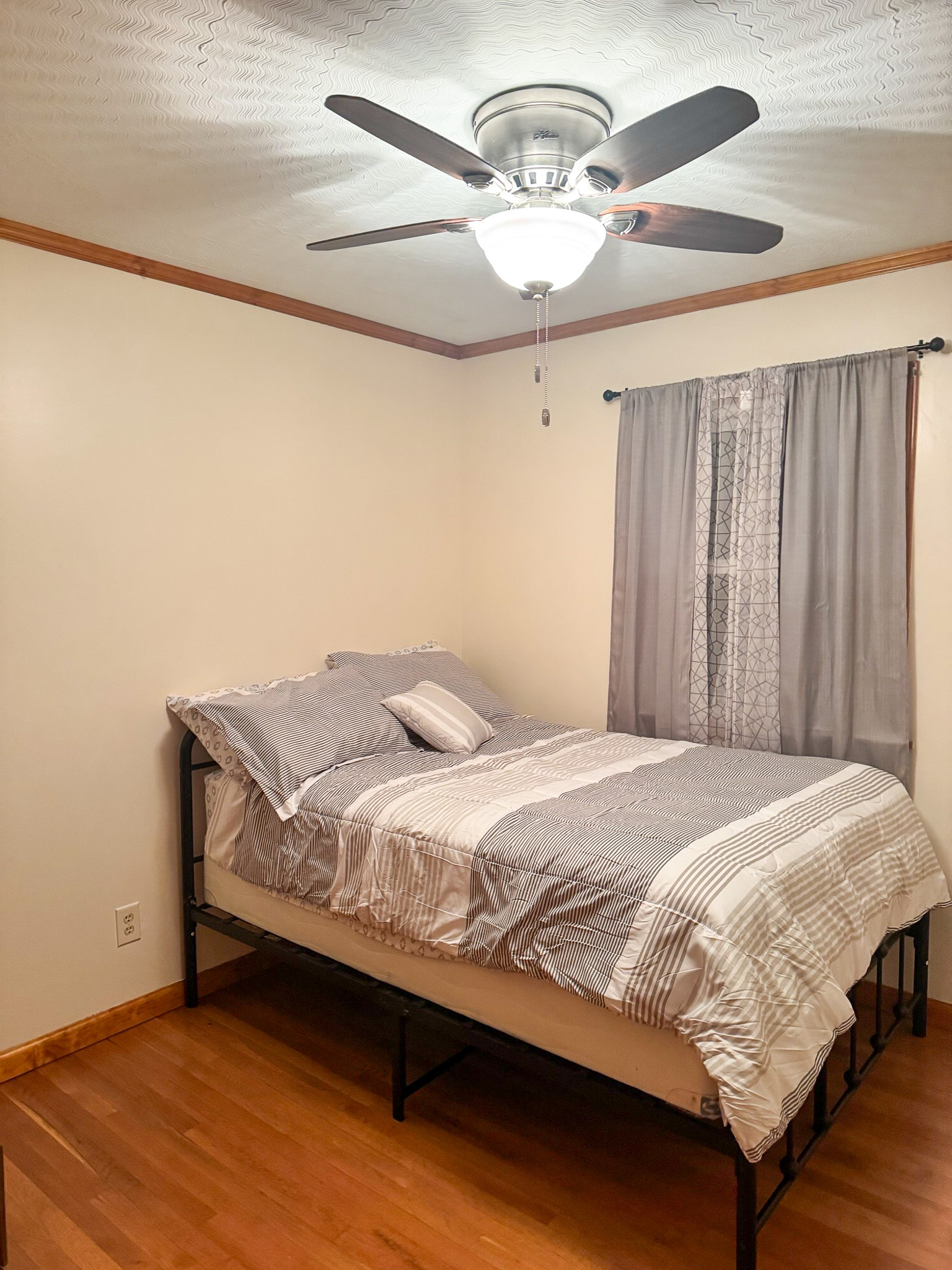 A bedroom with a neatly made bed in gray bedding, hardwood floors, light-colored walls, a ceiling fan with lights, and a window with gray curtains inside the home of Sponsored Residential Provider Pamela Hill in Austinville, Virginia.