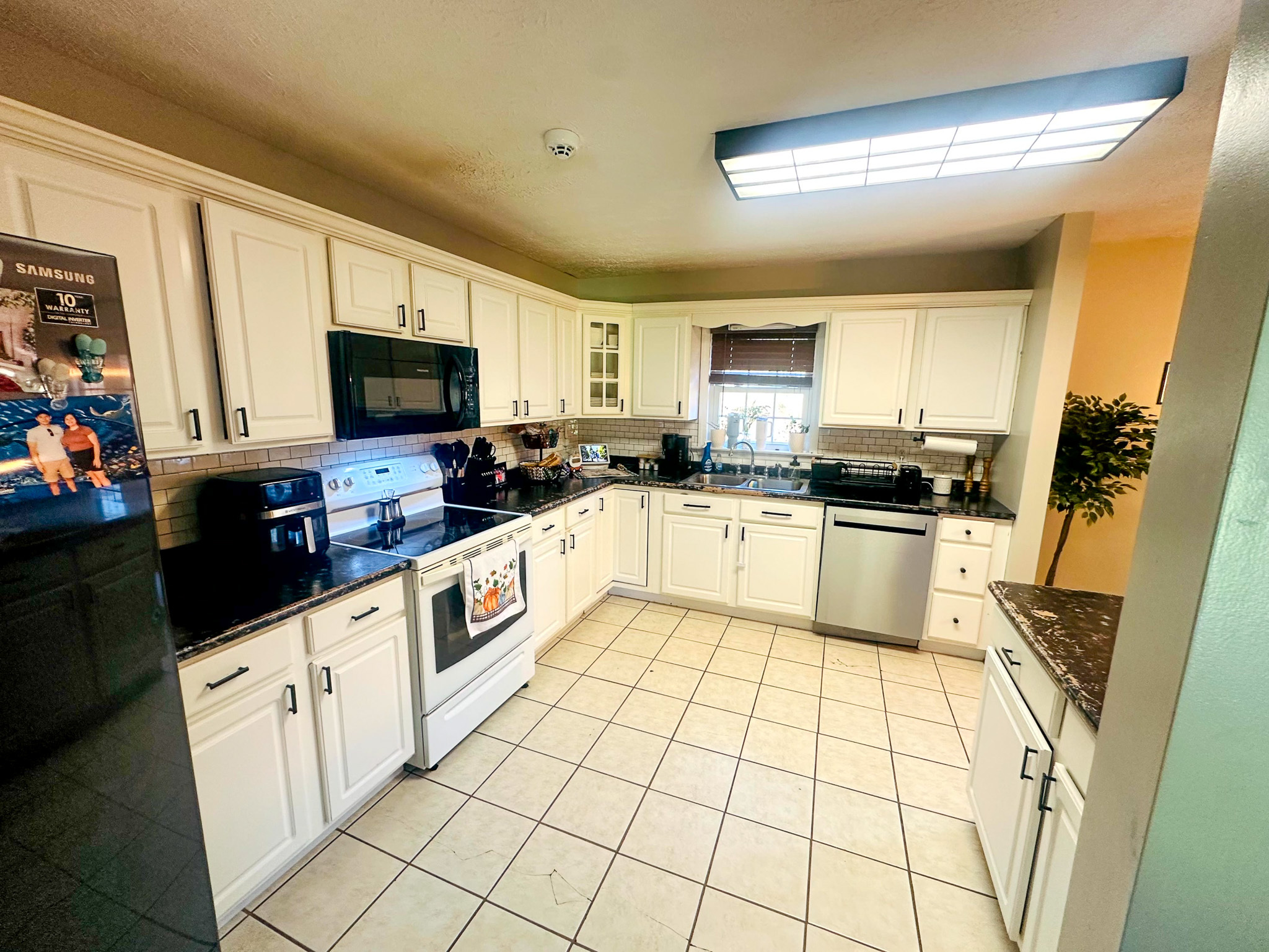 A bright U‑shaped kitchen with white cabinets, black countertops, tile flooring, and stainless‑steel appliances beneath a large overhead light inside the home of Sponsored Residential Provider Jayda Utt in Galax, Virginia.