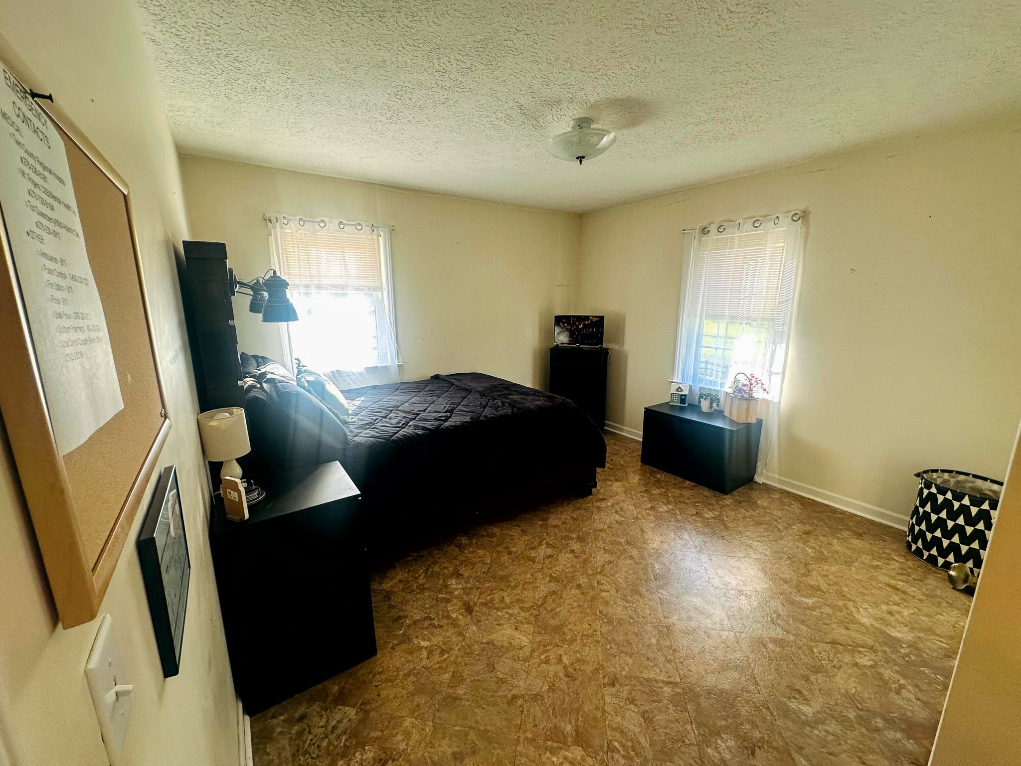 A spacious bedroom with light walls, two windows with sheer curtains, a dark bed and matching furniture, and patterned flooring under soft ceiling lighting inside the home of Sponsored Residential Provider Jayda Utt in Galax, Virginia.