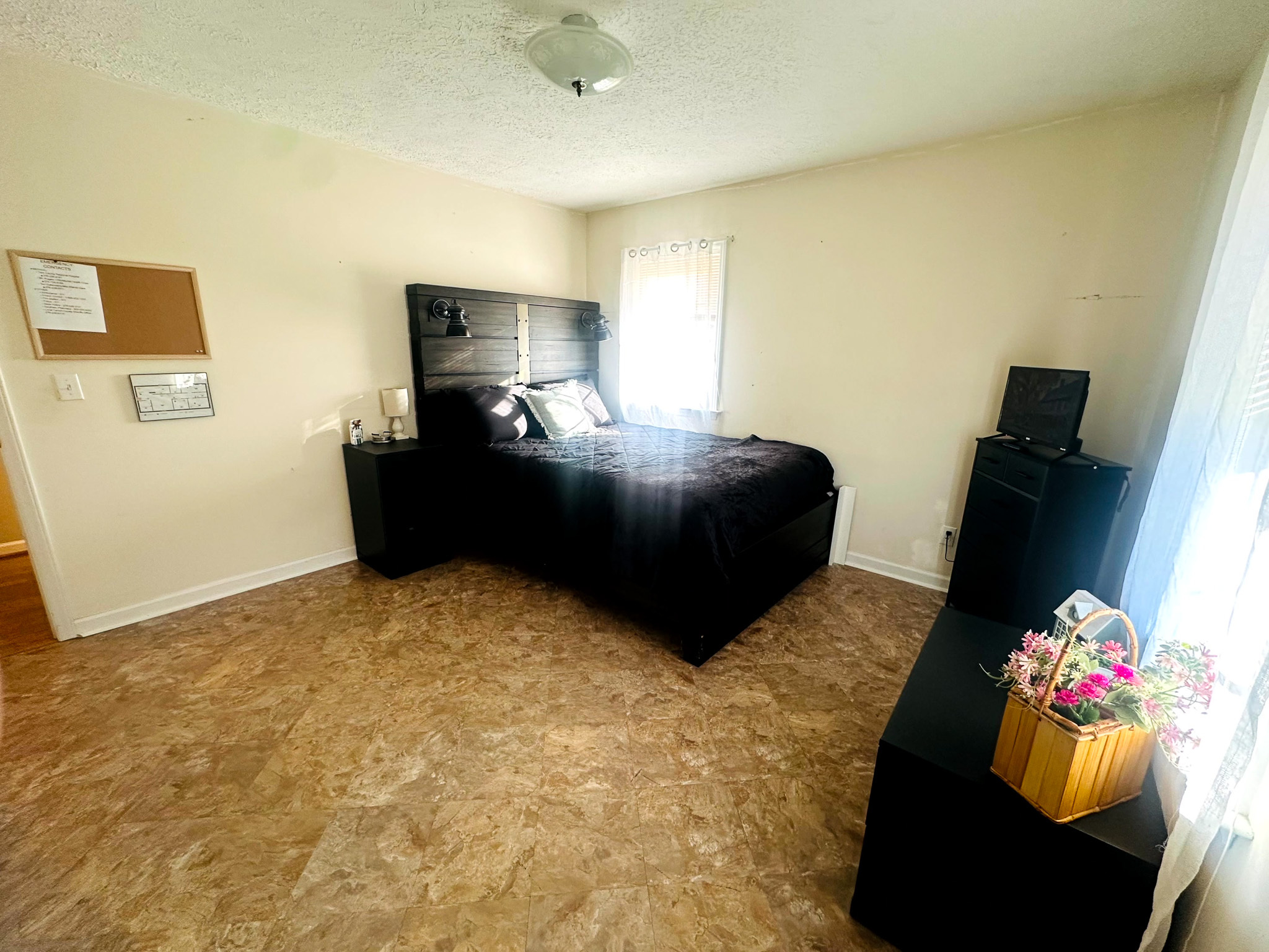 A bright bedroom with a black bed, matching black furniture, beige walls, and a light brown tile-patterned floor inside the home of Sponsored Residential Provider Jayda Utt in Galax, Virginia.