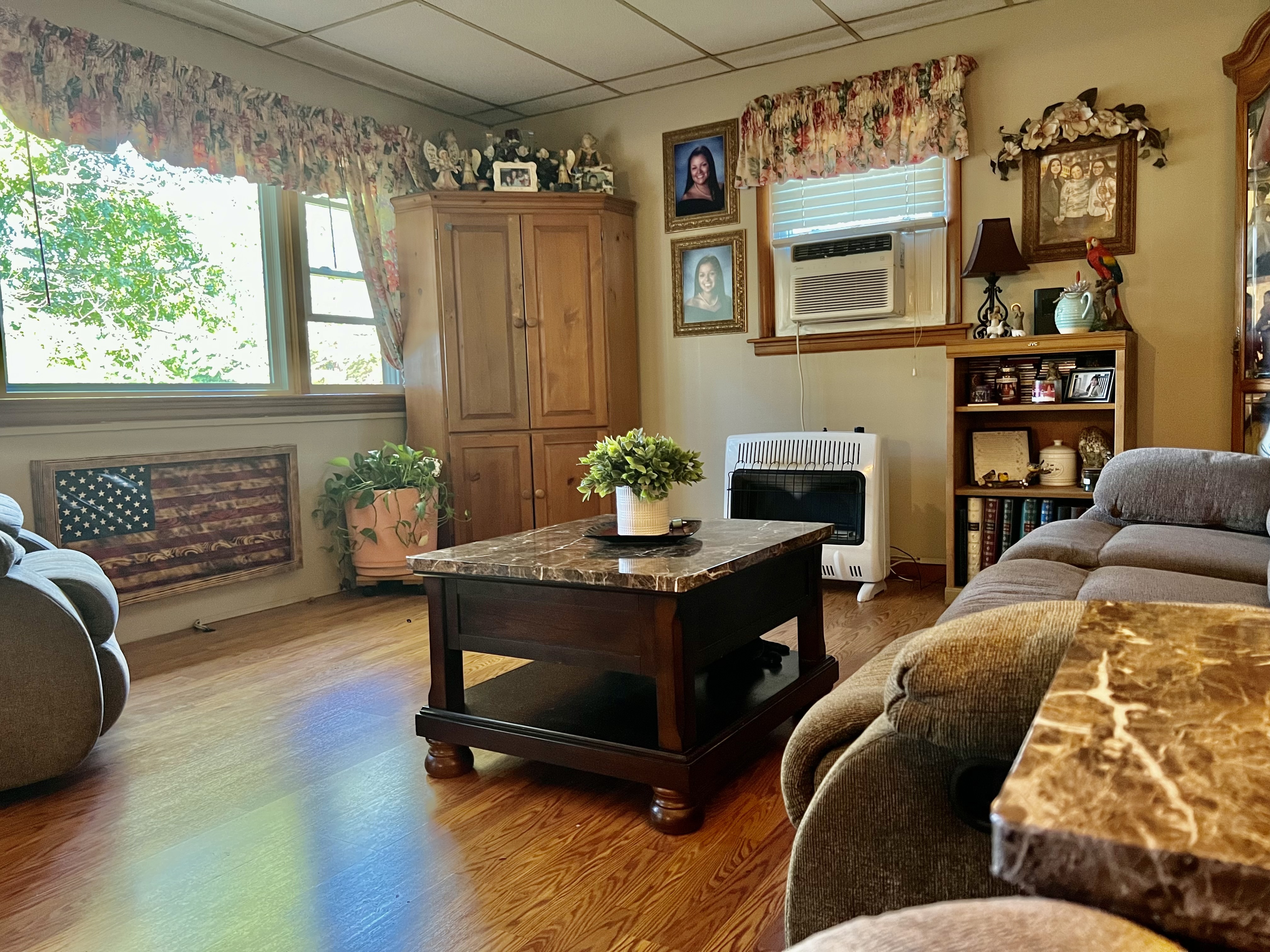 A bright living room with a marble‑top coffee table, reclining sofas, an air‑conditioning unit in the window, and wooden cabinets and shelves decorated with plants and framed photos inside the home of Sponsored Residential Providers Beth and Otis Fowler in Rustburg, Virginia.