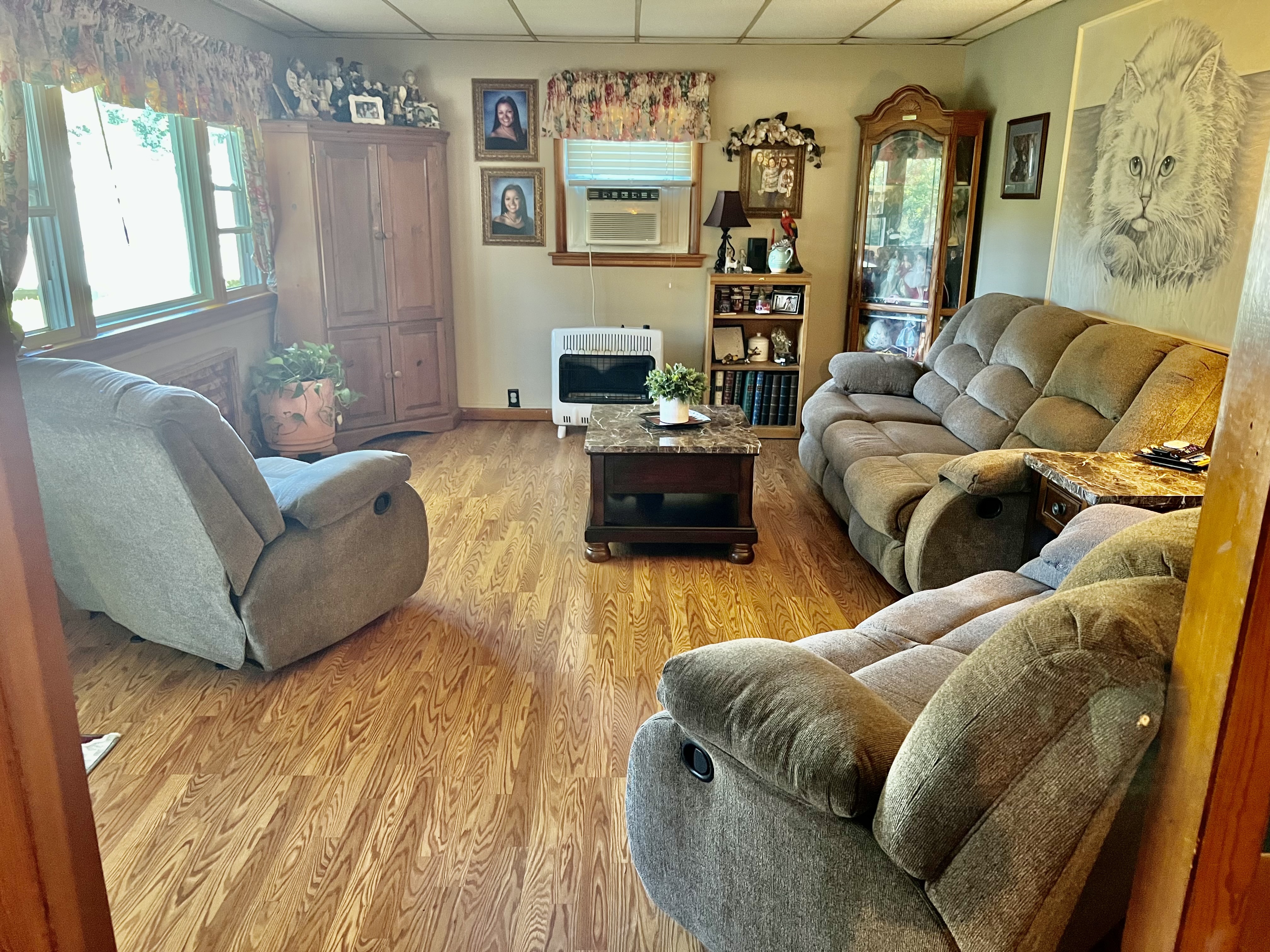 A cozy living room with reclining sofas, a wooden coffee table, and a corner cabinet, decorated with framed photos, plants, and a glass display cabinet inside the home of Sponsored Residential Providers Beth and Otis Fowler in Rustburg, Virginia.