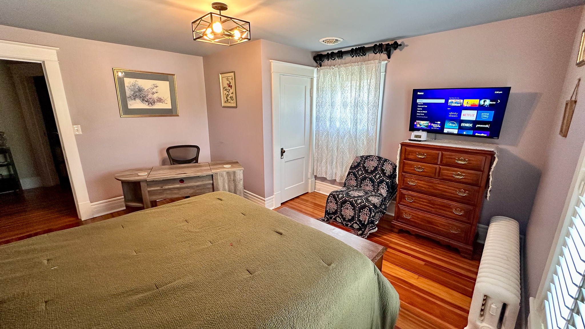 Bedroom with a green‑covered bed, corner desk and chair, patterned armchair, tall dresser with a wall‑mounted TV above it, and hardwood floors inside the home of Sponsored Residential Providers Jeff and Sarah McLane in Lynchburg, Virginia.
