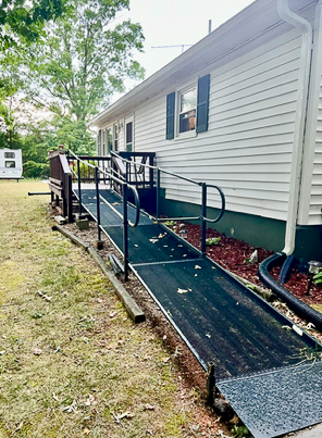 A metal accessibility ramp with railings runs alongside a white house with green shutters, set over mulch and leading to a small porch belonging to Sponsored Residential Providers Beth and Otis Fowler in Rustburg, Virginia.