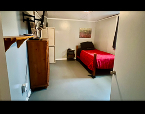 A simple bedroom with a single bed covered in a red blanket, light gray walls, wooden furniture, and a wall‑mounted TV inside the home of Sponsored Residential Providers Beth and Otis Fowler in Rustburg, Virginia.