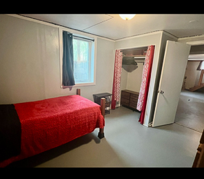 A bedroom with a single bed covered in a red and black blanket, a window with dark curtains, and an open closet area with a red-patterned curtain inside the home of Sponsored Residential Providers Beth and Otis Fowler in Rustburg, Virginia.